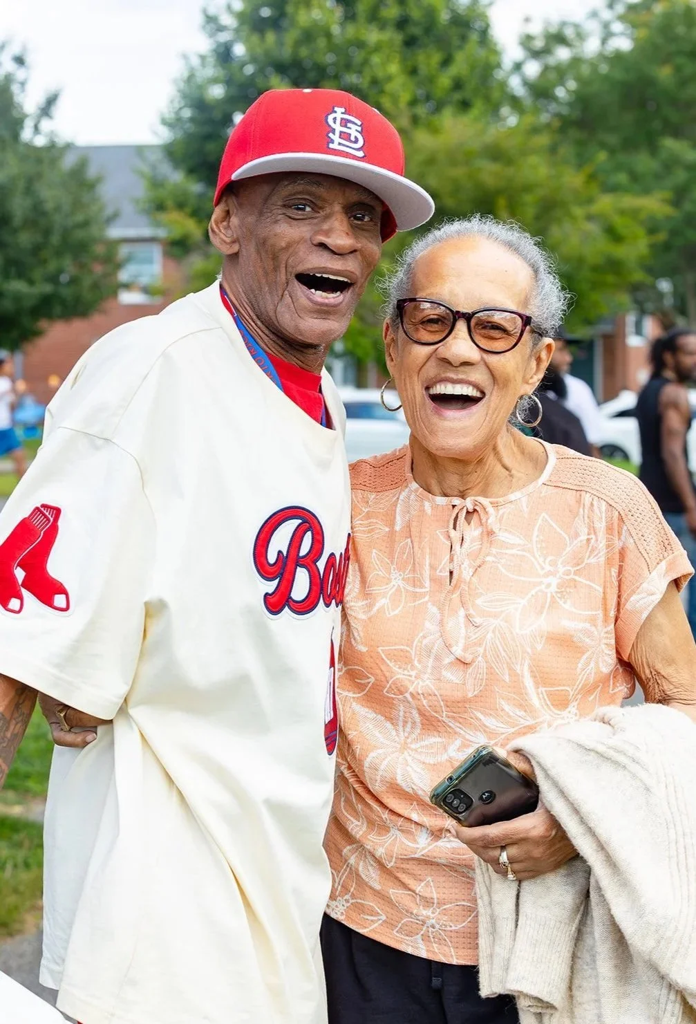 A man wearing a baseball jersey and a red cap, and a woman wearing glasses and a floral top, are smiling and enjoying a moment outdoors among other people and trees.
