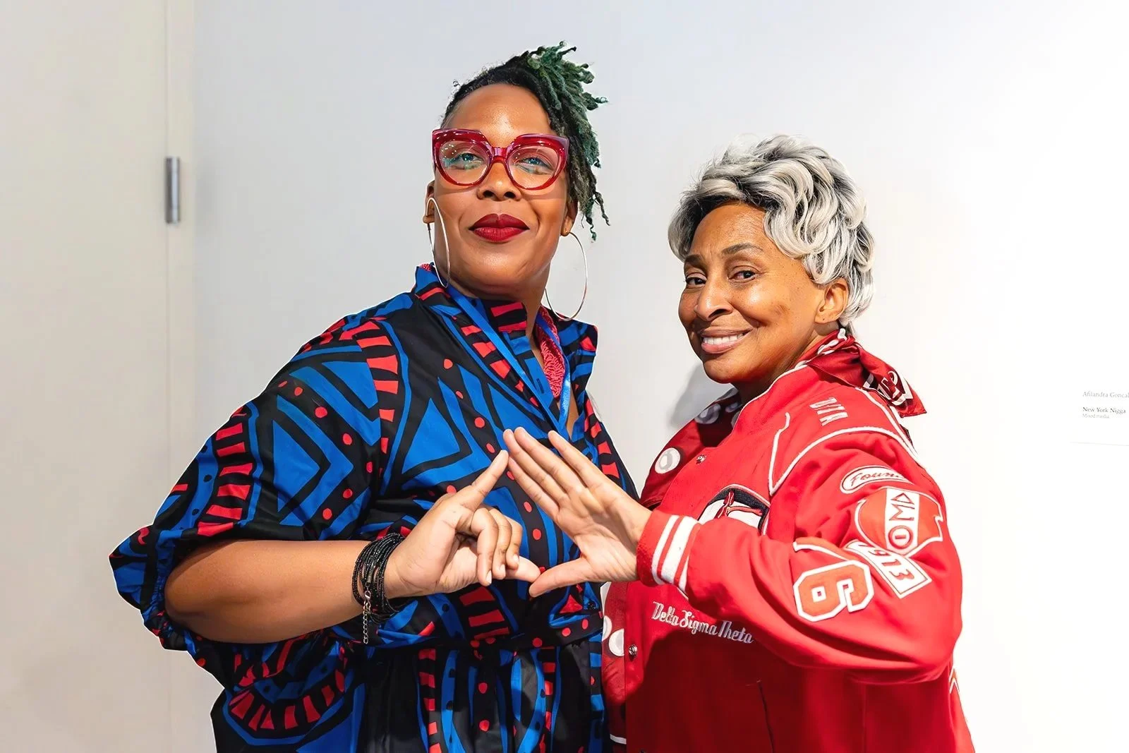 Two women standing close together, making a heart shape with their hands. The woman on the left has short locks with green tips, wearing large red glasses, a colorful patterned dress, and hoop earrings. The woman on the right has short gray hair, wea