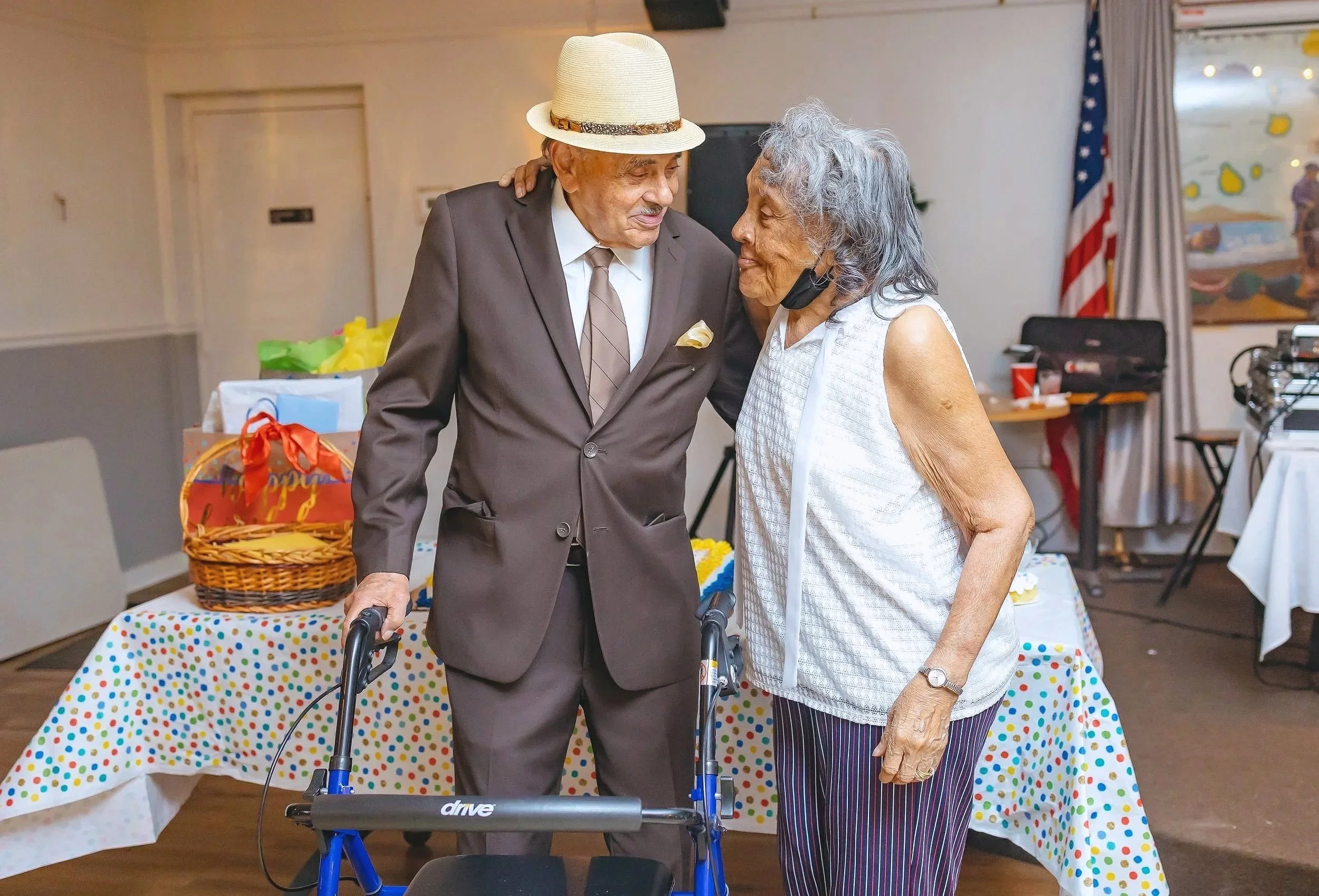 An elderly man in a brown suit and a woman in a white sleeveless top share a tender moment.