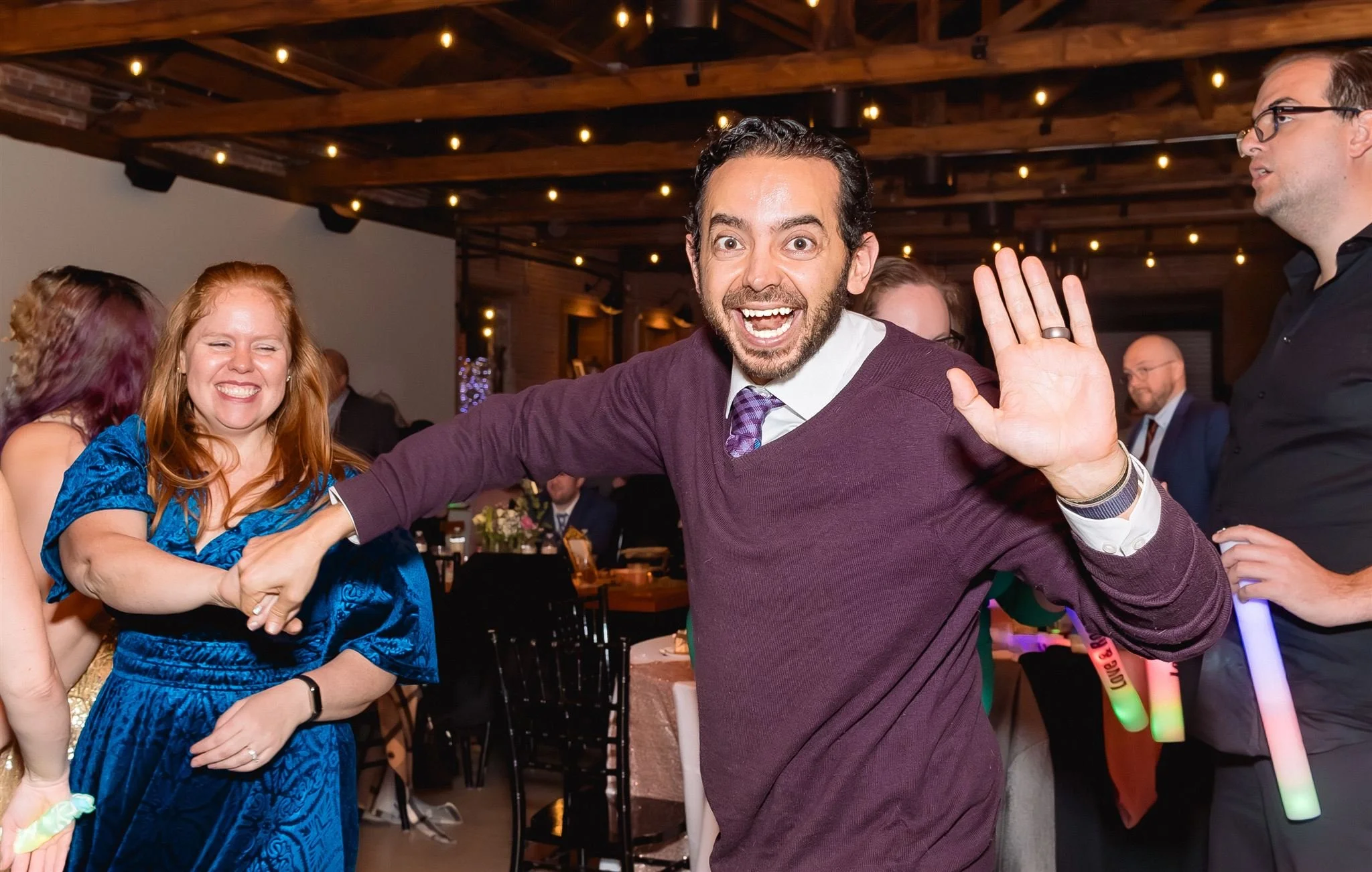 Group of people celebrating indoors with joyful expressions, some dancing and holding glow sticks, in a warmly lit venue with wooden ceiling beams.