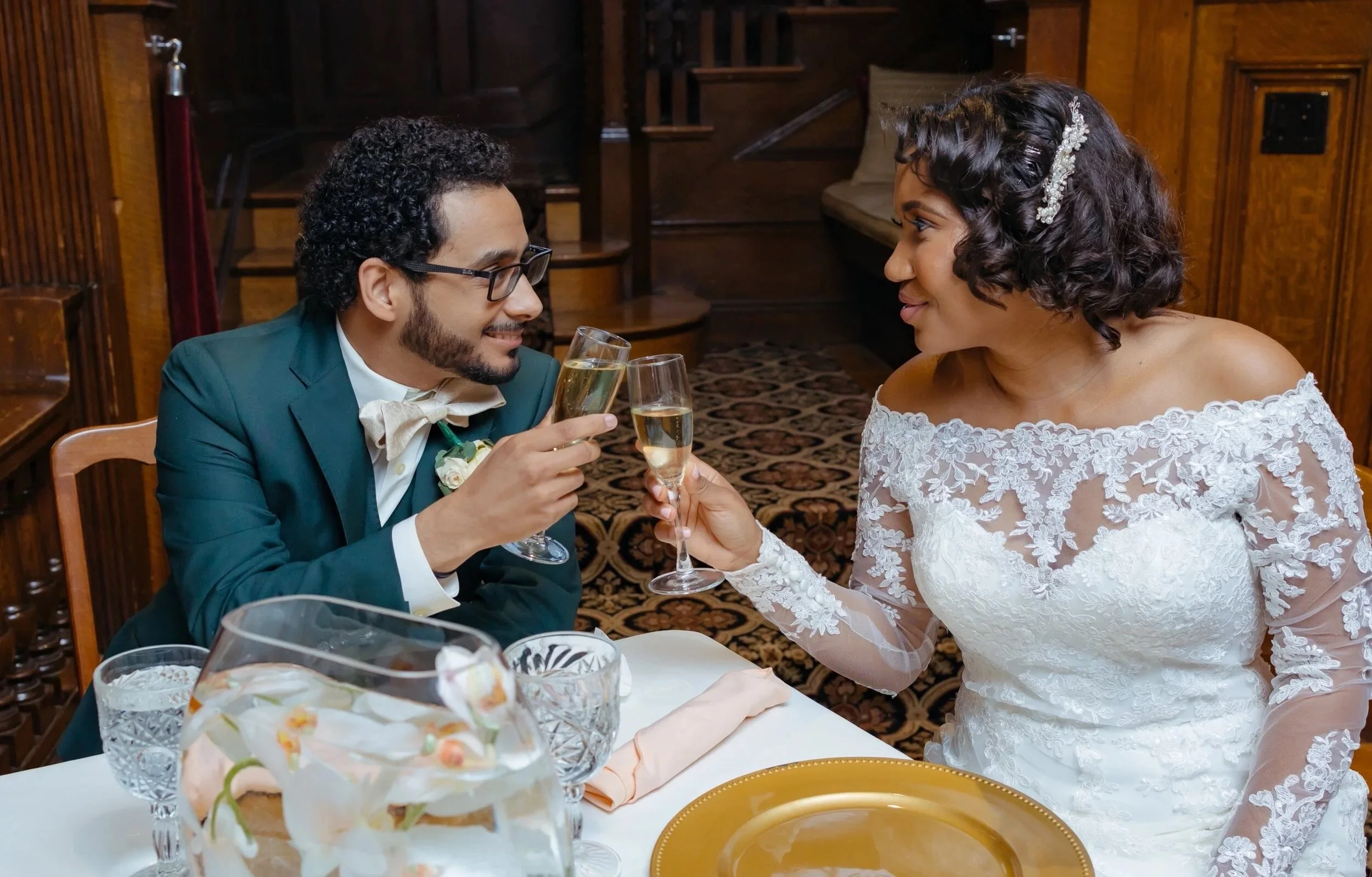 A couple celebrating at a wedding reception, clinking champagne glasses, with the bride in a lace wedding dress and the groom in a suit and bowtie, sitting at a decorated table in an elegant wooden venue.