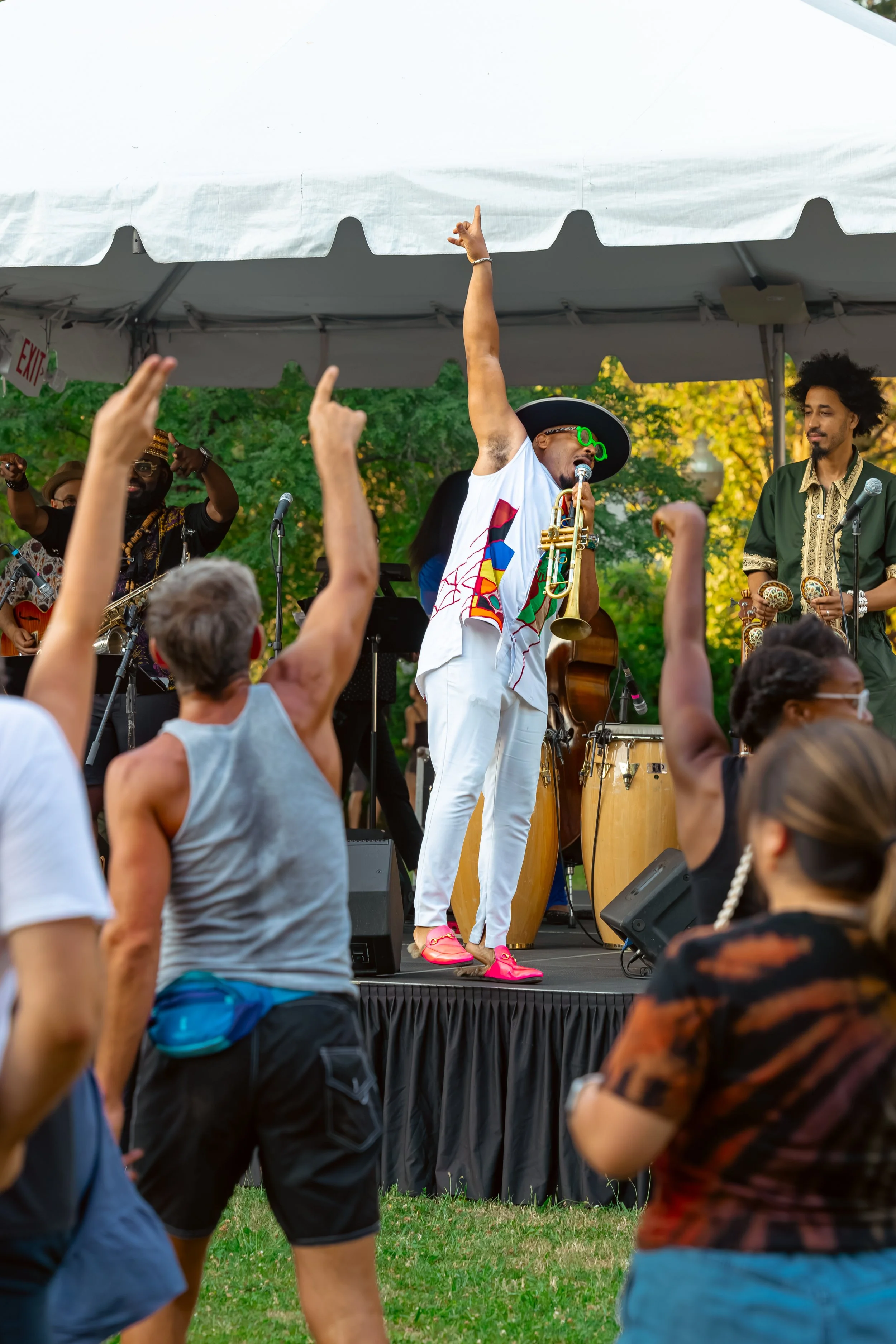 Musicians performing on an outdoor stage at a festival or concert, playing instruments and engaging with the audience. Audience members are dancing and raising their hands in front of the stage, set against a background of green trees.