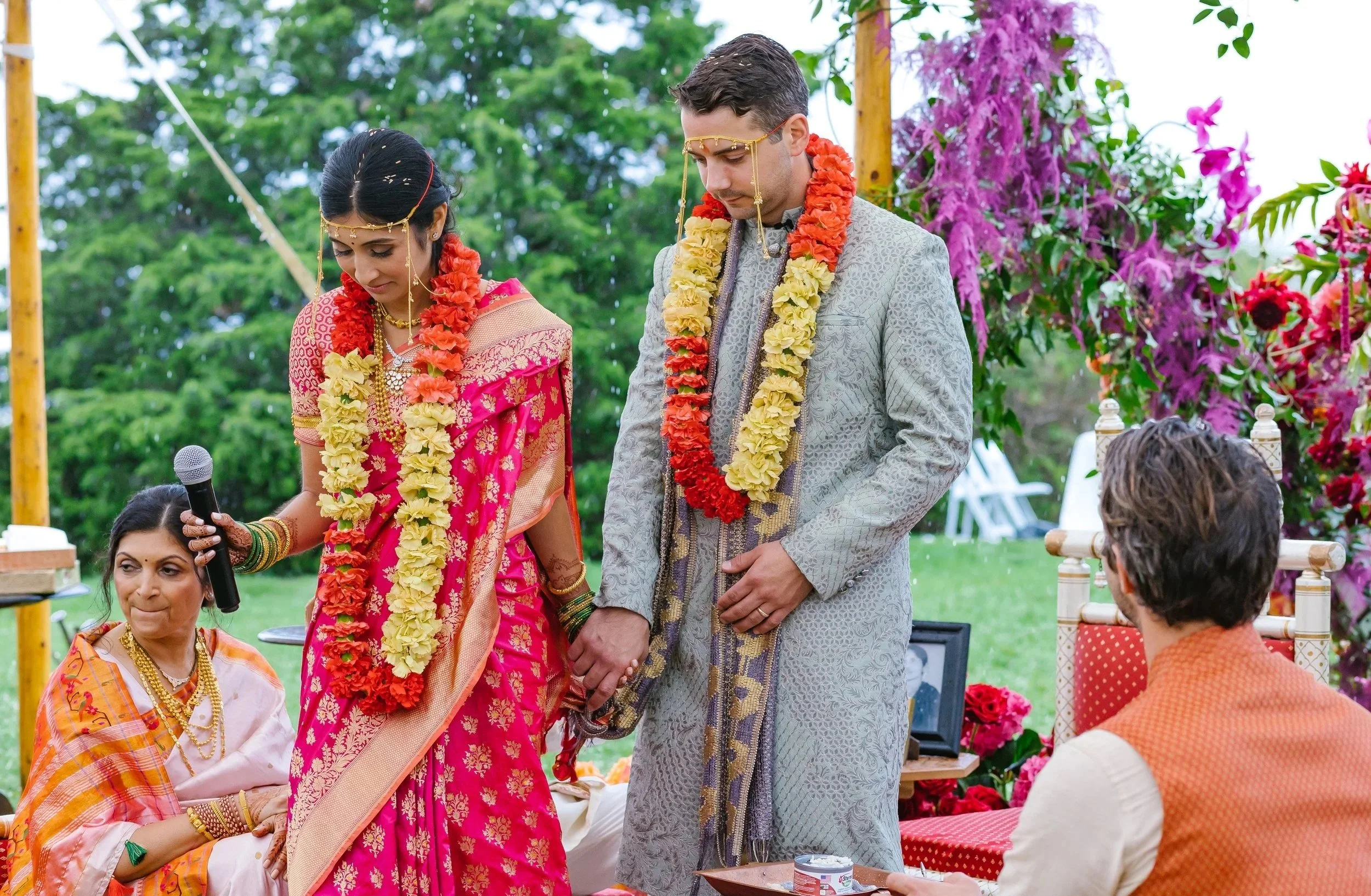 Indian wedding ceremony with bride and groom holding hands, surrounded by family, outdoors with greenery and colorful flowers.