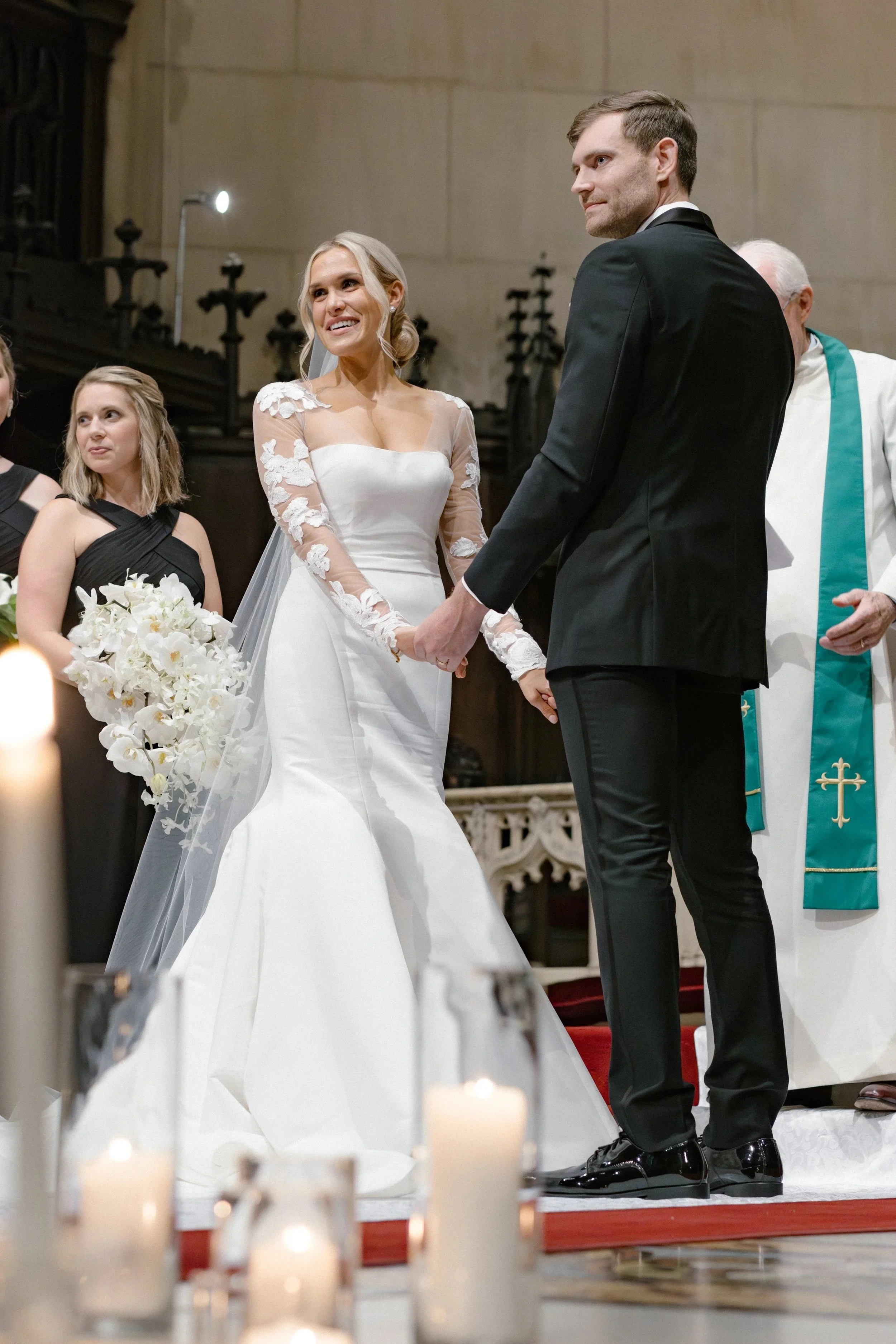A couple gets married in a church, holding hands and smiling at each other. The bride is in a white wedding dress with lace sleeves and is holding a bouquet of white flowers. The groom is in a black tuxedo. Other people and candles are in the backgro