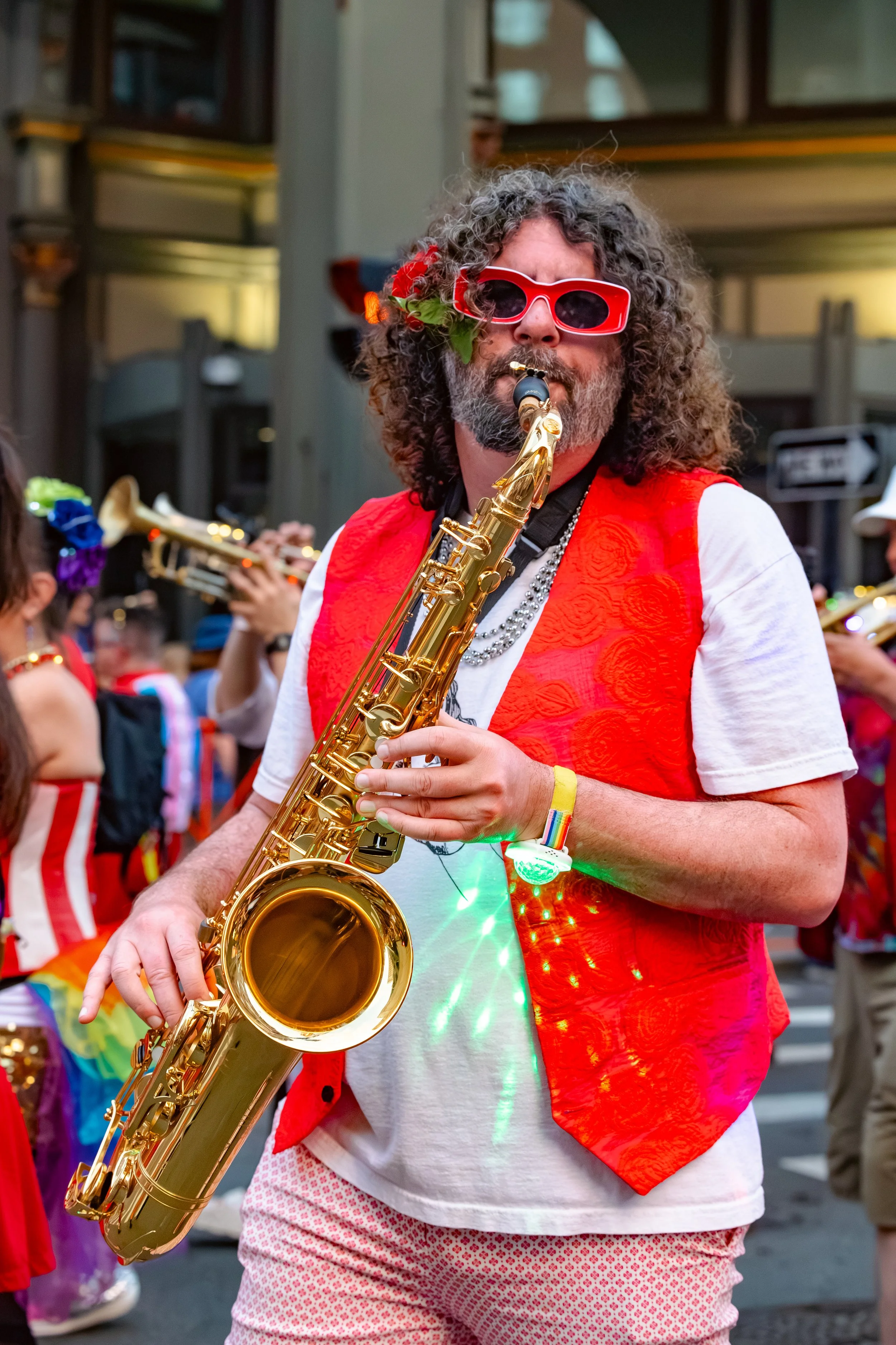 A man with curly hair, wearing red sunglasses, and a red vest, playing a gold saxophone during a parade or festival.