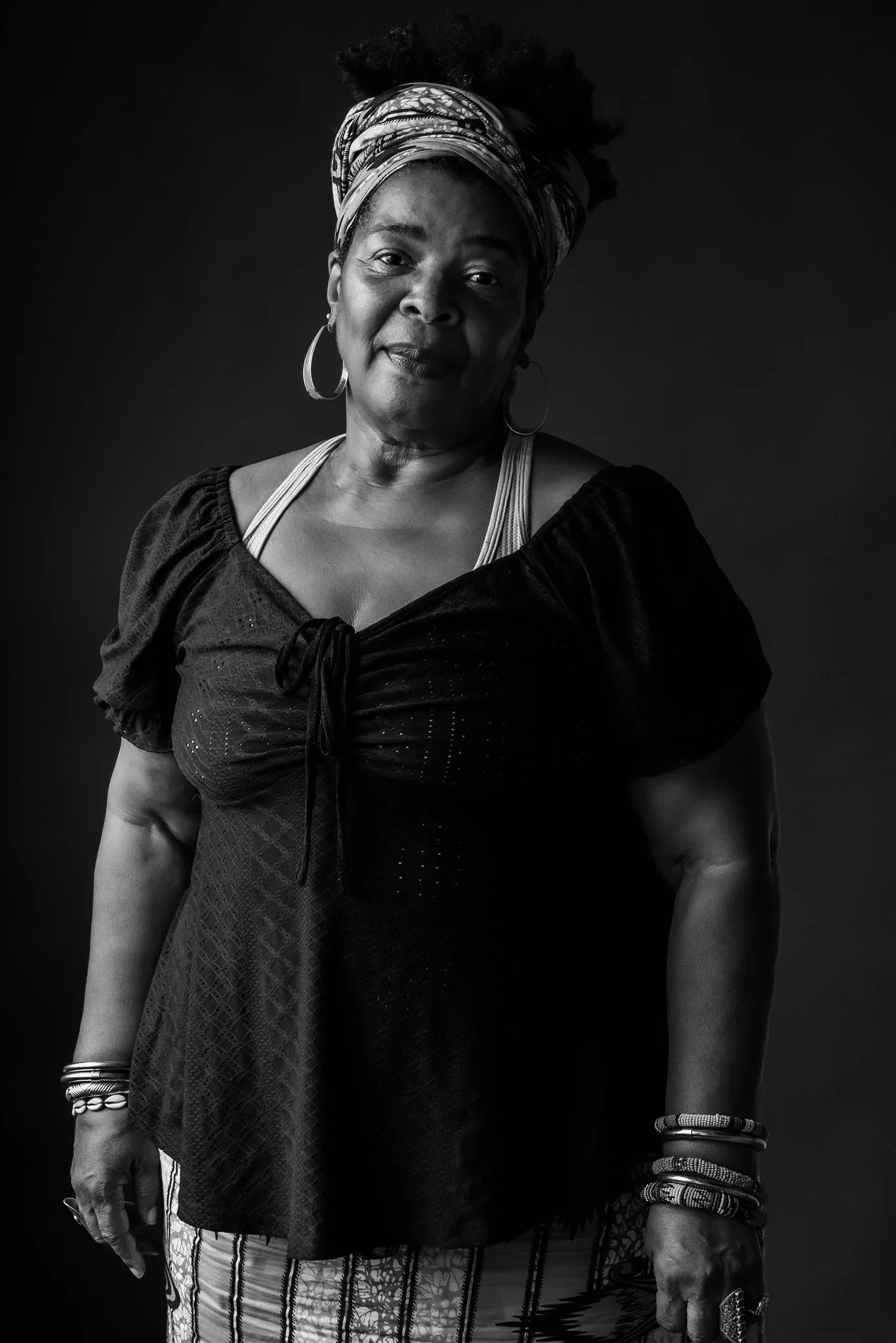 A black and white portrait of an older woman wearing a headwrap, earrings, and multiple bracelets, standing against a dark background.