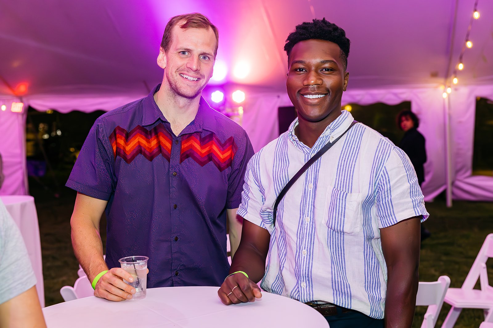 Two men smiling at a social event under a lit canopy, one holding a drink, with other people in the background.