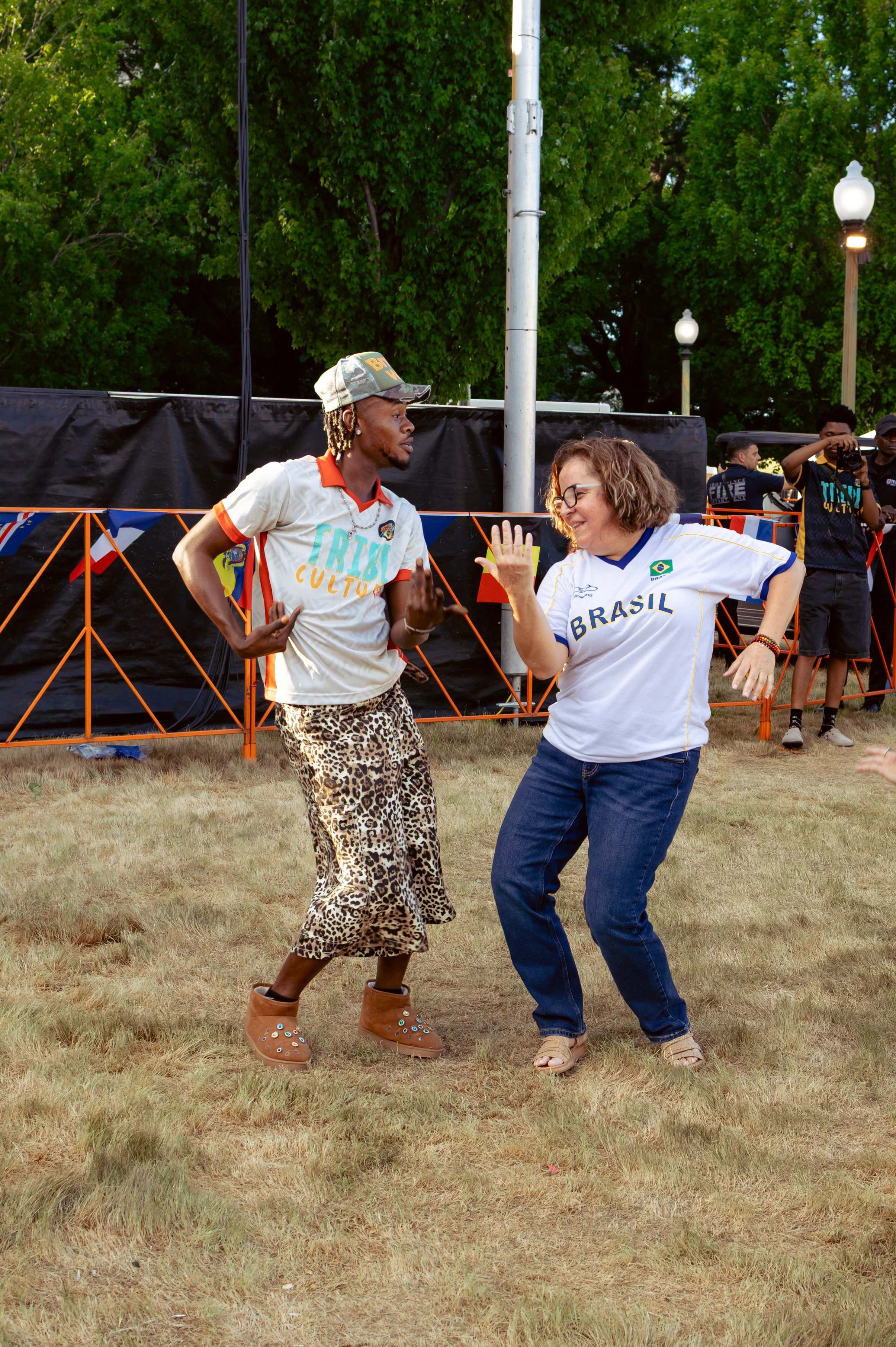 Two women dancing outdoors in a grassy area with trees, a black fence, and street lamps in the background. One woman wears a white shirt with Brazil's flag, glasses, and casual pants. The other woman has leopard print pants, a cap, and a colorful shi