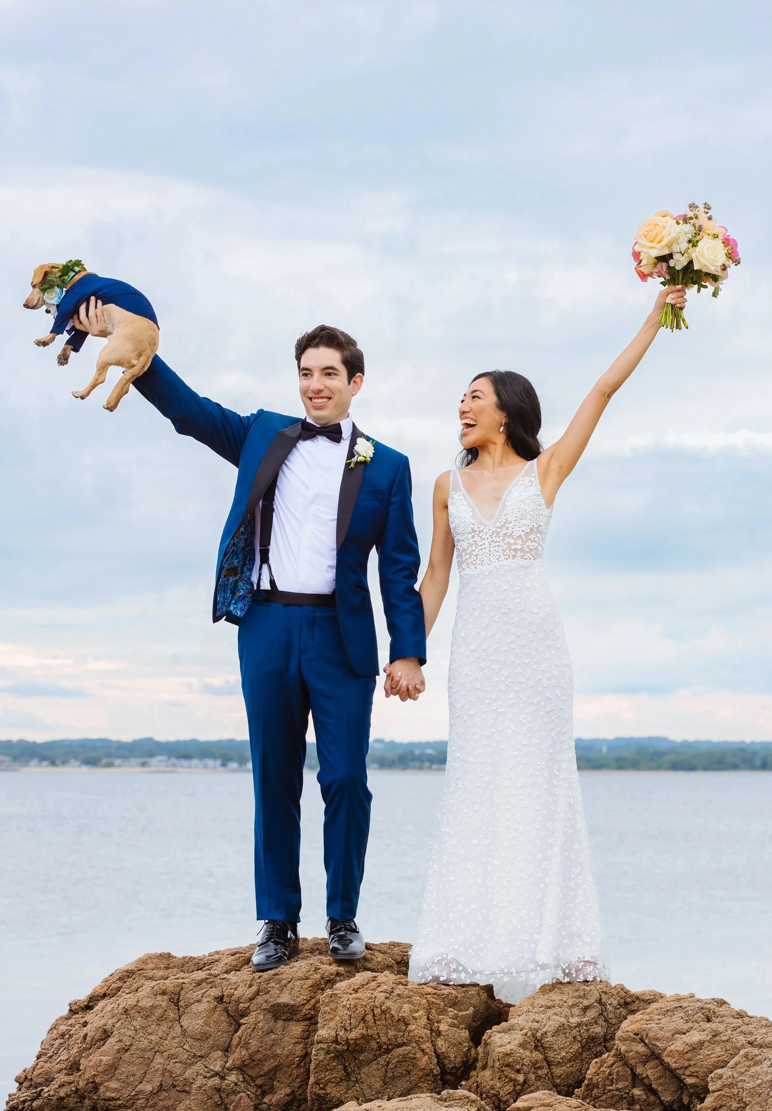 A newlywed couple standing on rocks by a body of water, holding hands, with the bride raising her bouquet and the groom lifting a small dog dressed in a blue outfit, all smiling happily.