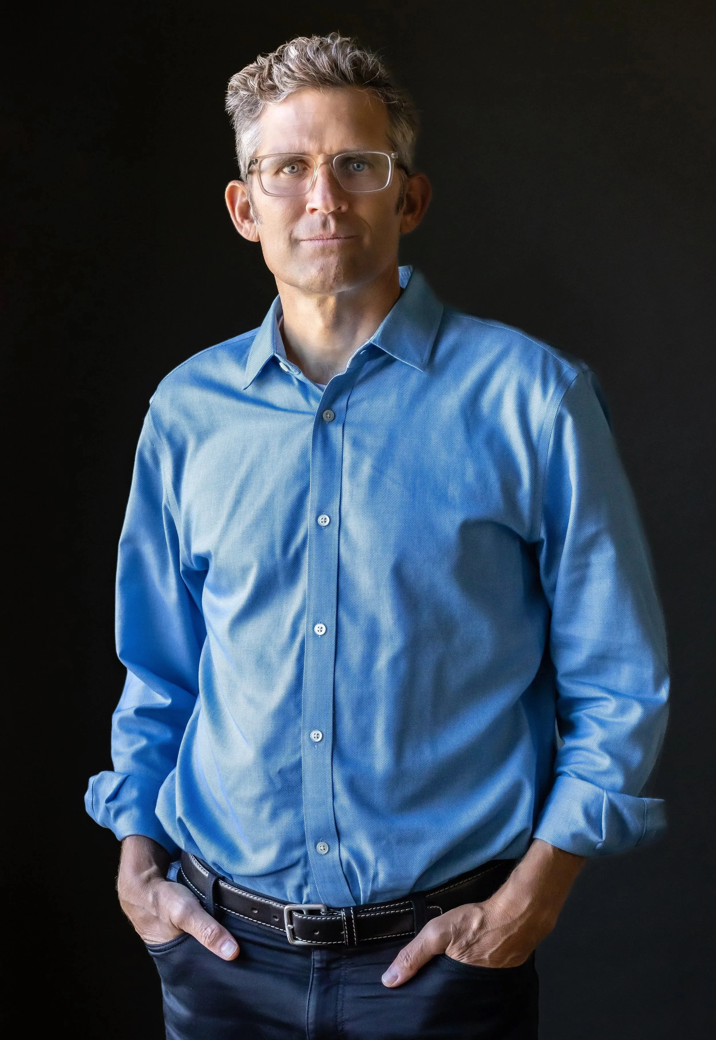 A middle-aged man with glasses and gray hair, wearing a blue button-up shirt and black pants, standing against a dark background.
