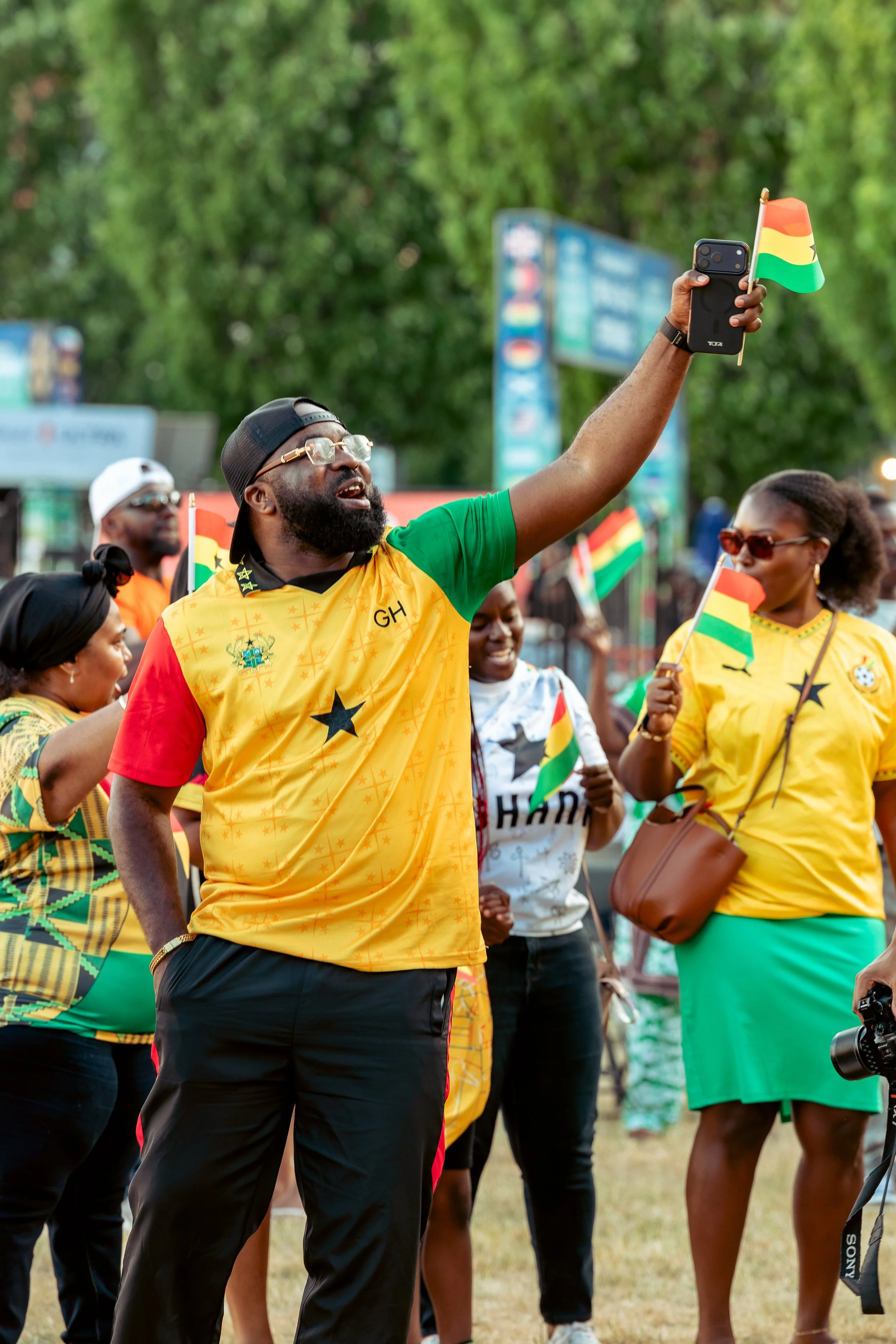 A group of people celebrating Ghana's Independence Day outdoors. They are wearing colorful clothing with Ghanaian flags and symbols, some holding small flags. One man in the center is taking a selfie, holding a smartphone with a Ghanaian flag attache