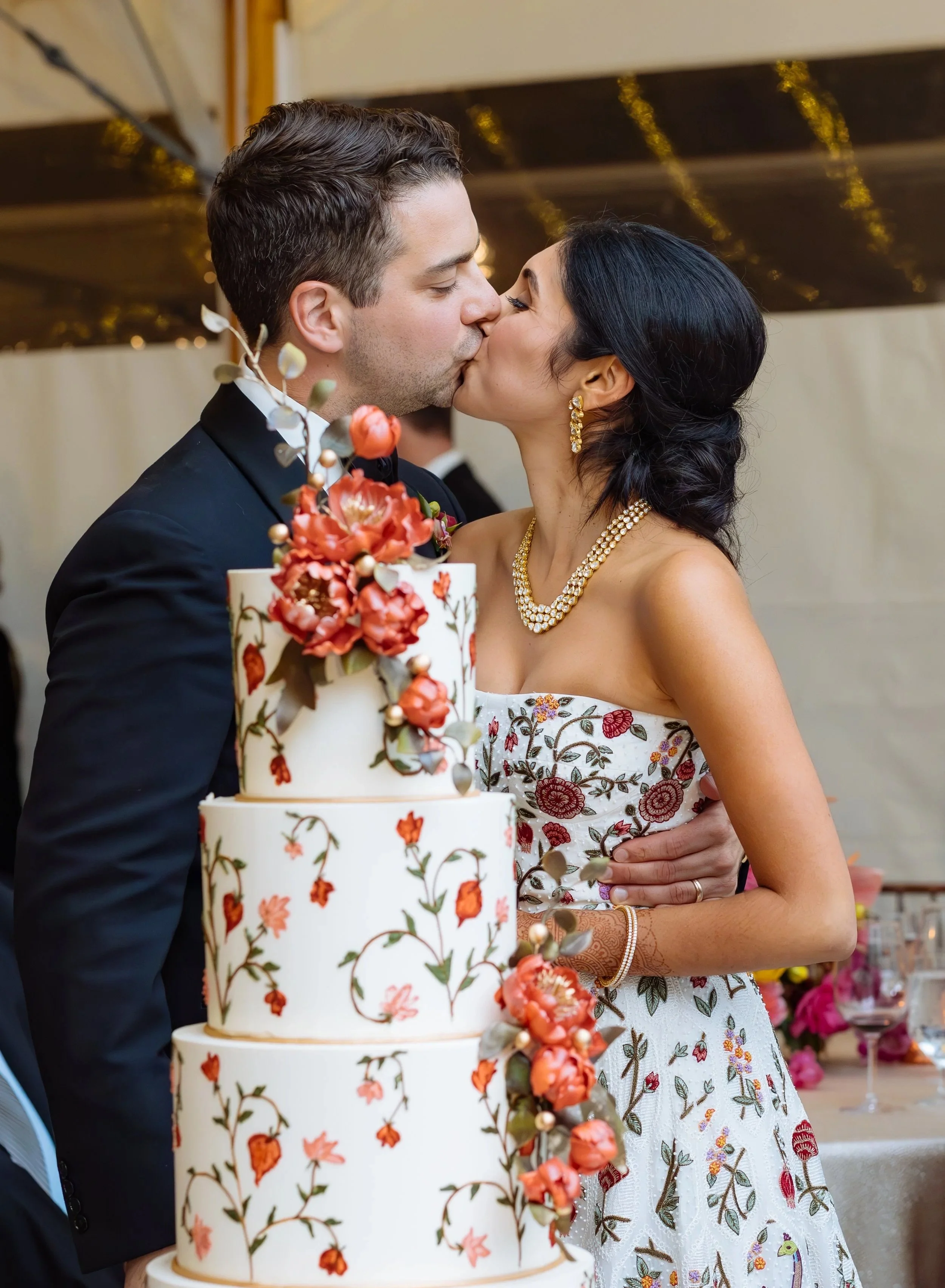 A bride and groom kissing at their wedding reception with a large floral wedding cake in front of them.