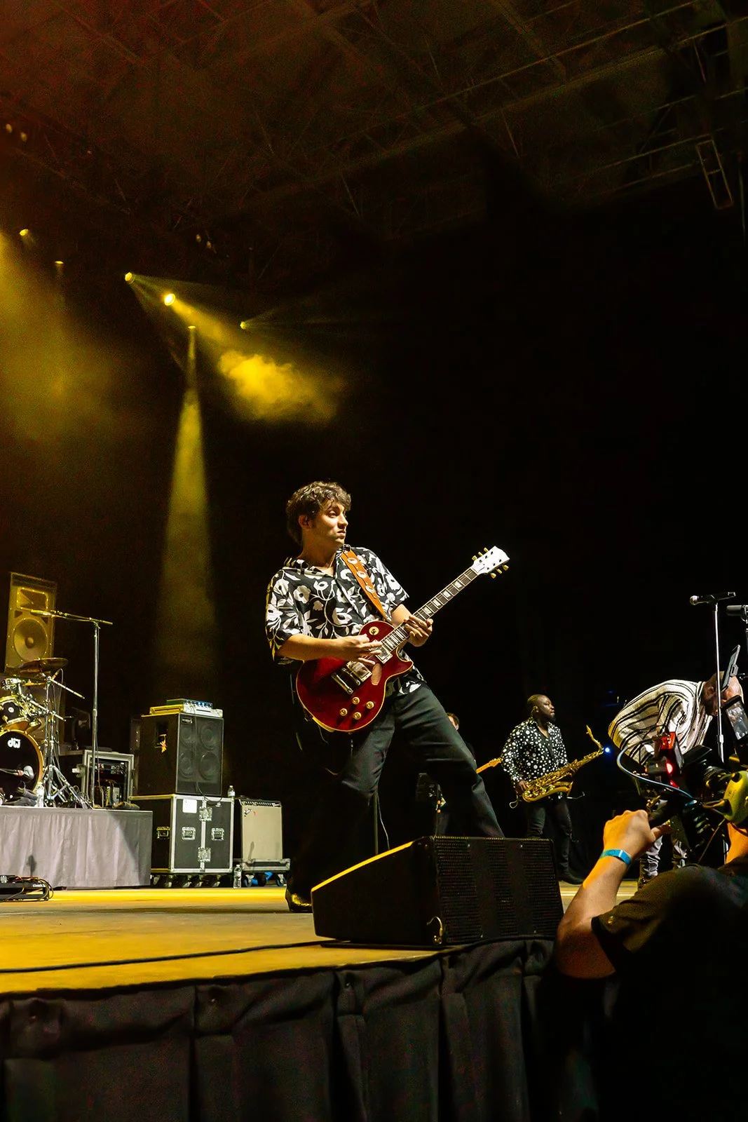 A musician playing an electric guitar on stage during a concert, with band members and musical equipment in the background, and stage lights illuminating the scene.