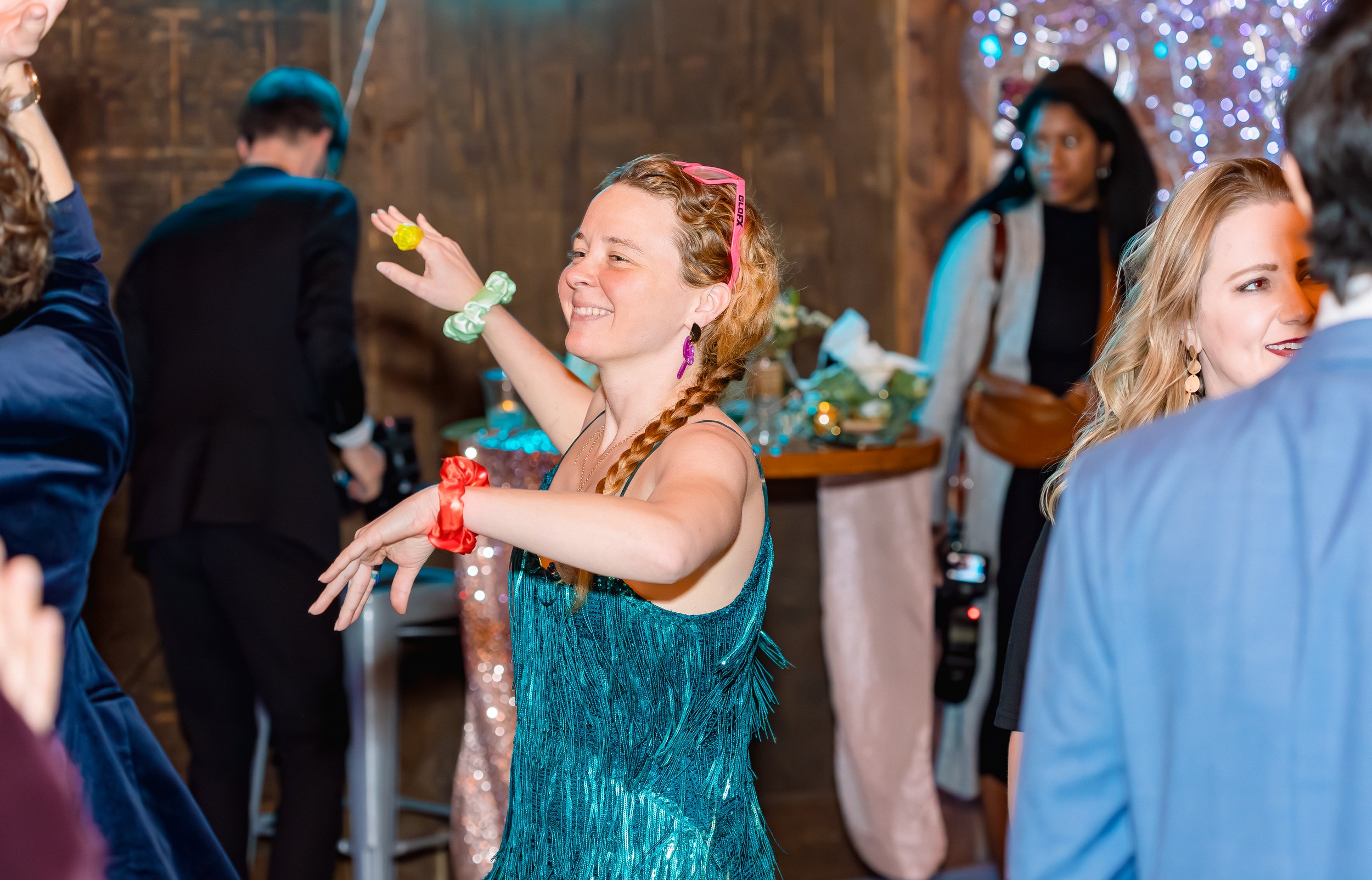 Woman dancing at a party, smiling, wearing a teal dress with fringe, pink sunglasses on her head, and colorful scrunchies on her wrists.