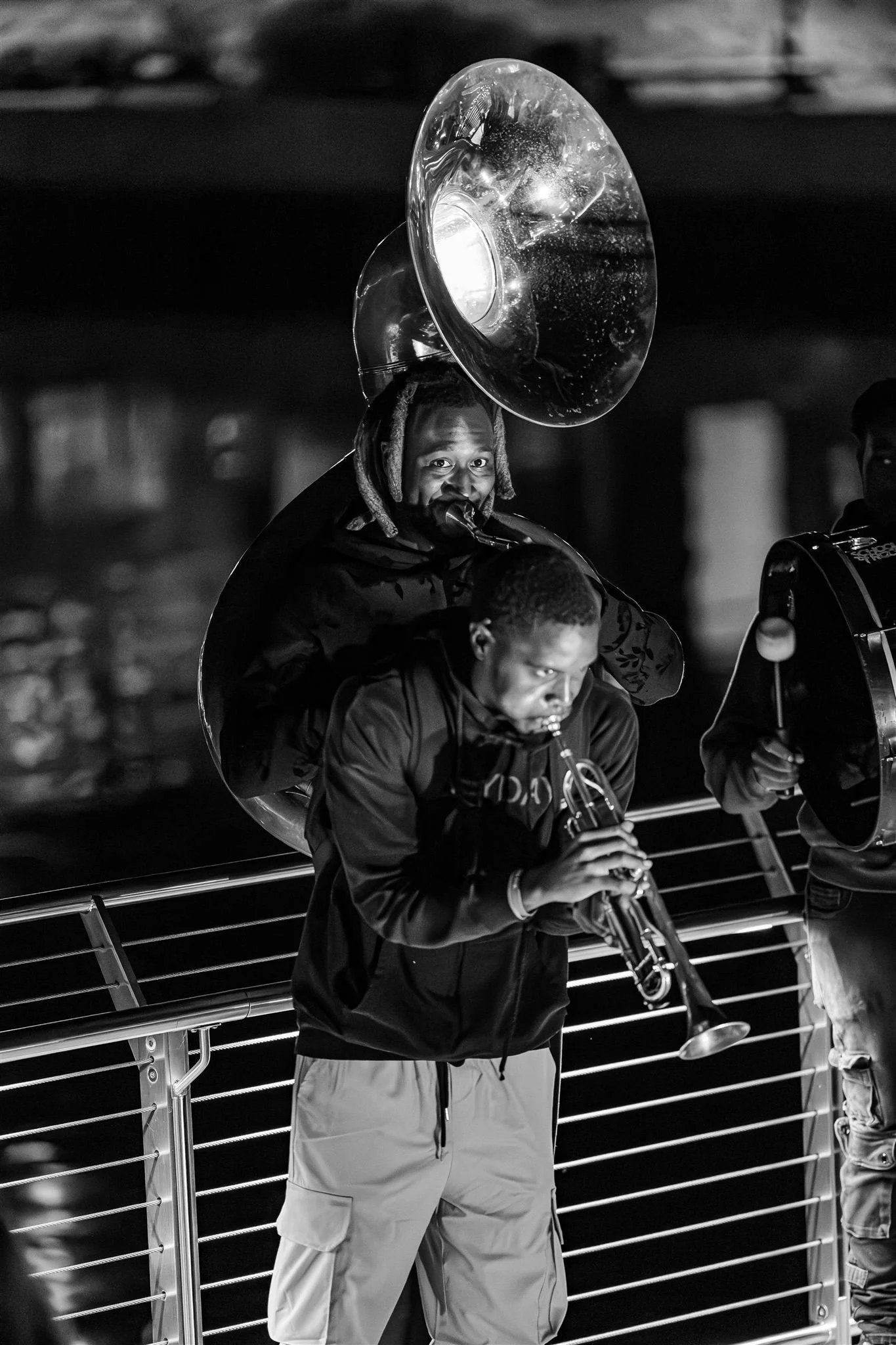 Two musicians playing brass instruments on an outdoor stage at night, one with a sousaphone and the other with a trumpet.