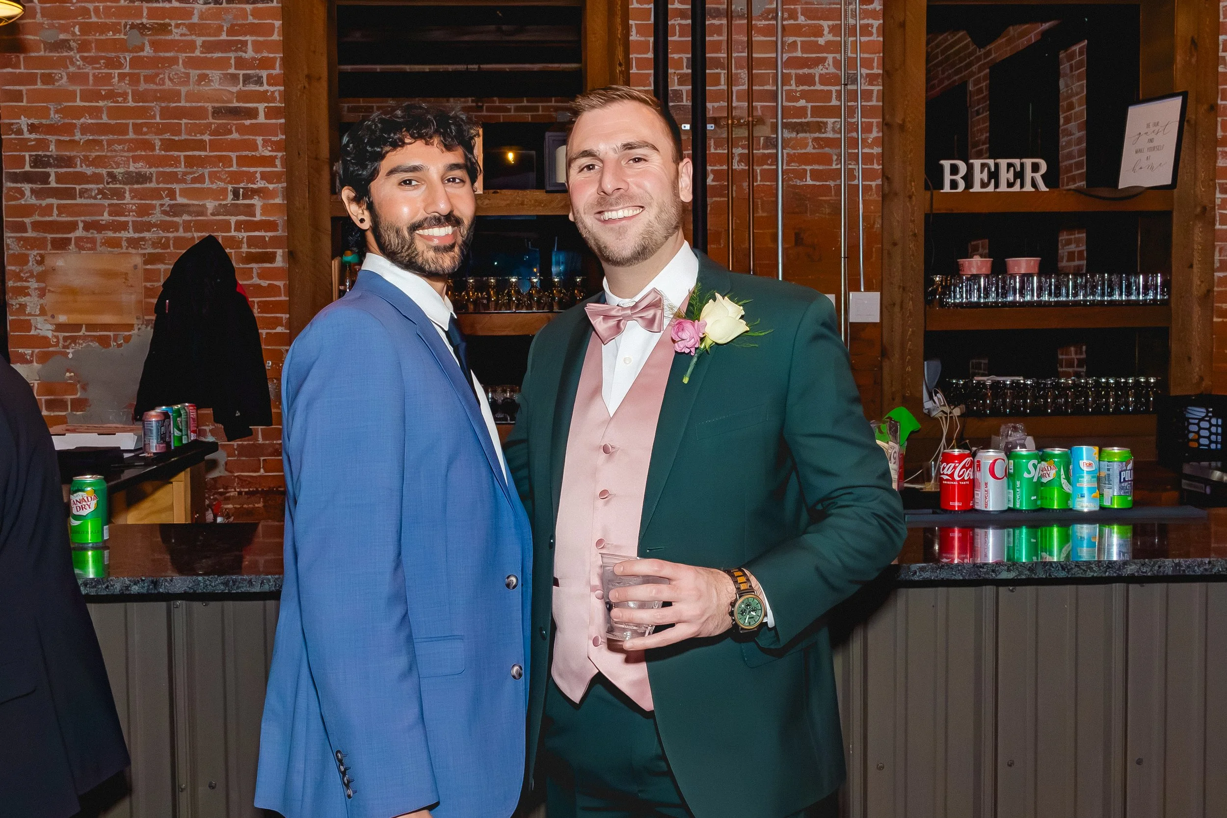Two men in suits at a bar, one wearing a blue suit and the other wearing a green suit with a pink bow tie and boutonniere, smiling and holding a drink, in a brick-walled venue with a bar and soda cans in the background.