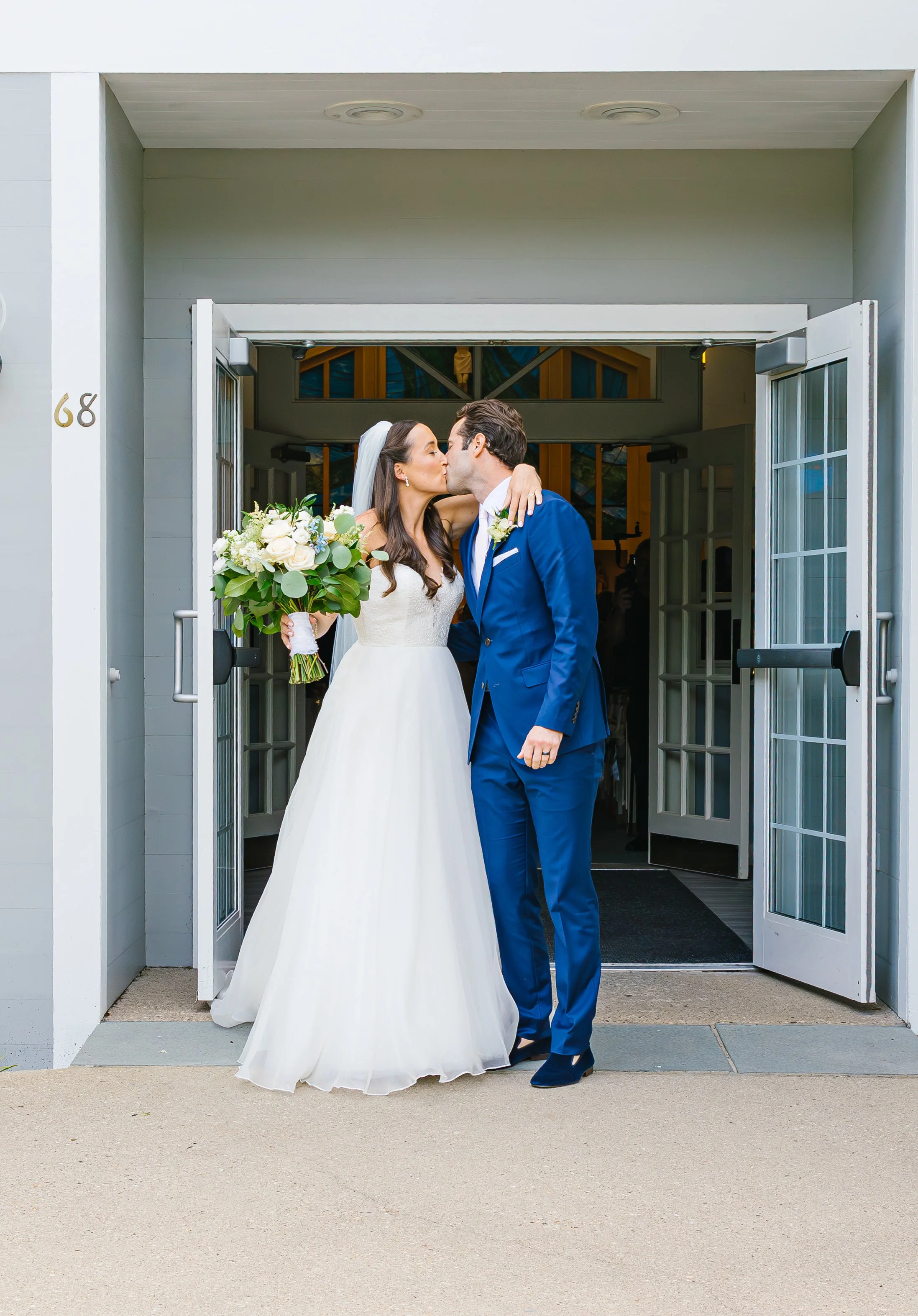 Bride and groom sharing a kiss outside the church after their wedding, with the bride holding a bouquet of white flowers and greenery.