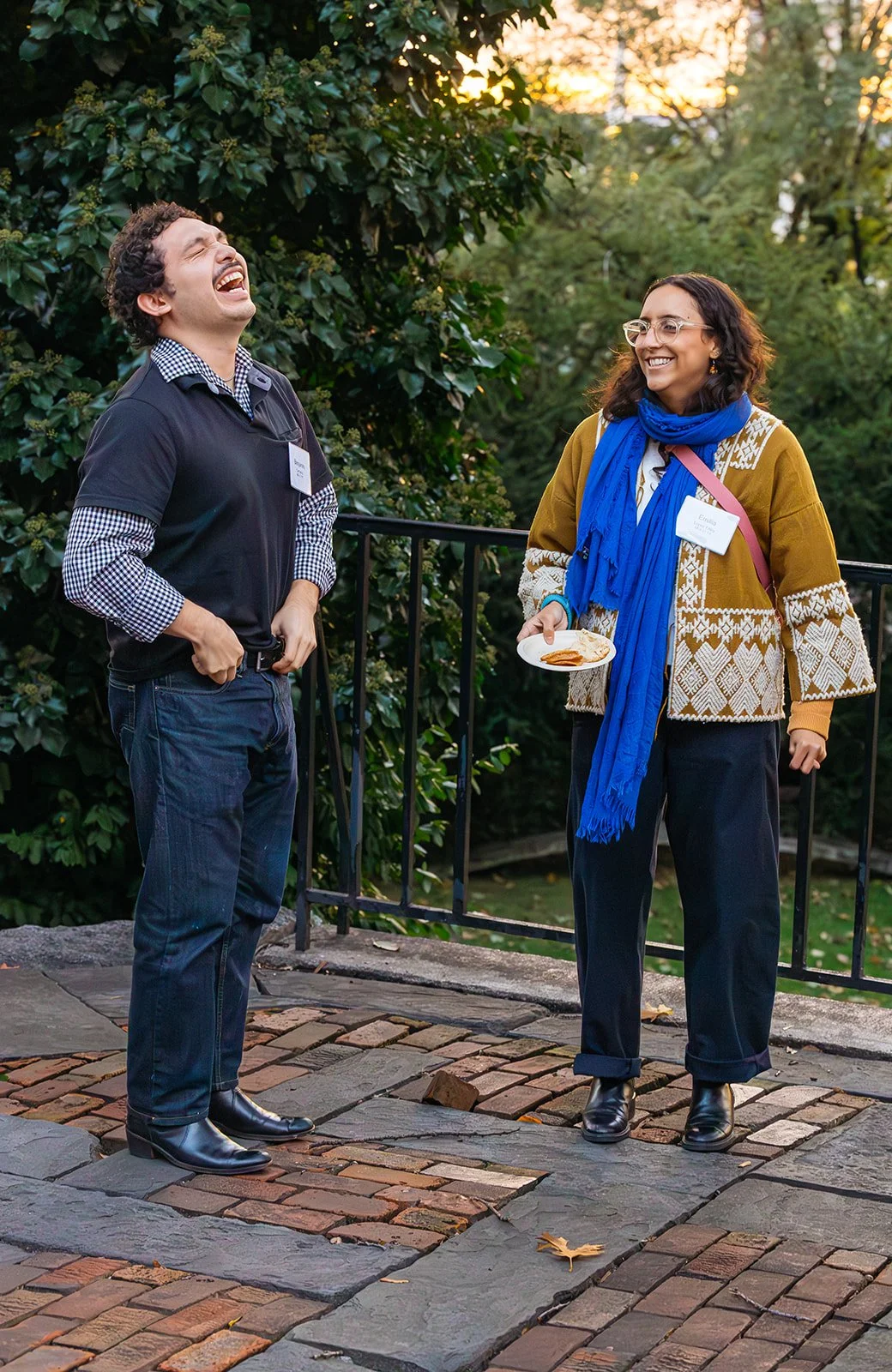 Two people standing outdoors near a black railing, smiling and laughing. The man has curly hair, wearing a black vest over a checkered shirt, jeans, and black boots. The woman has glasses, wavy hair, a colorful patterned sweater, a bright blue scarf,