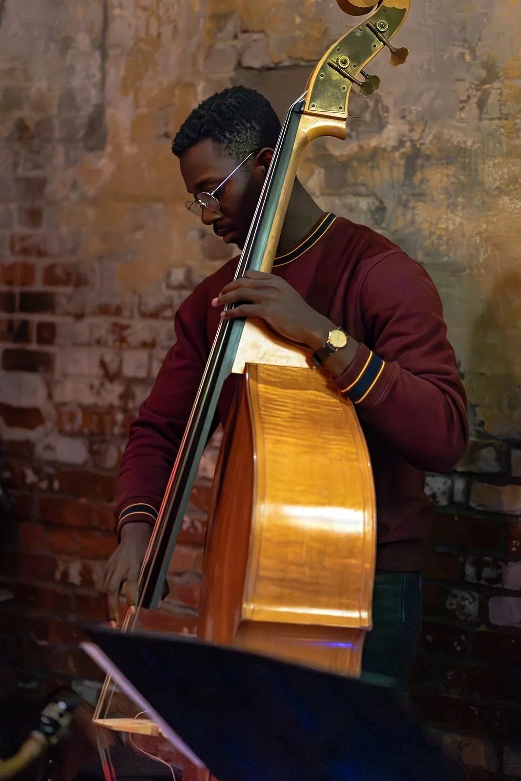 A man wearing glasses and a burgundy sweater playing a double bass against a brick wall background.