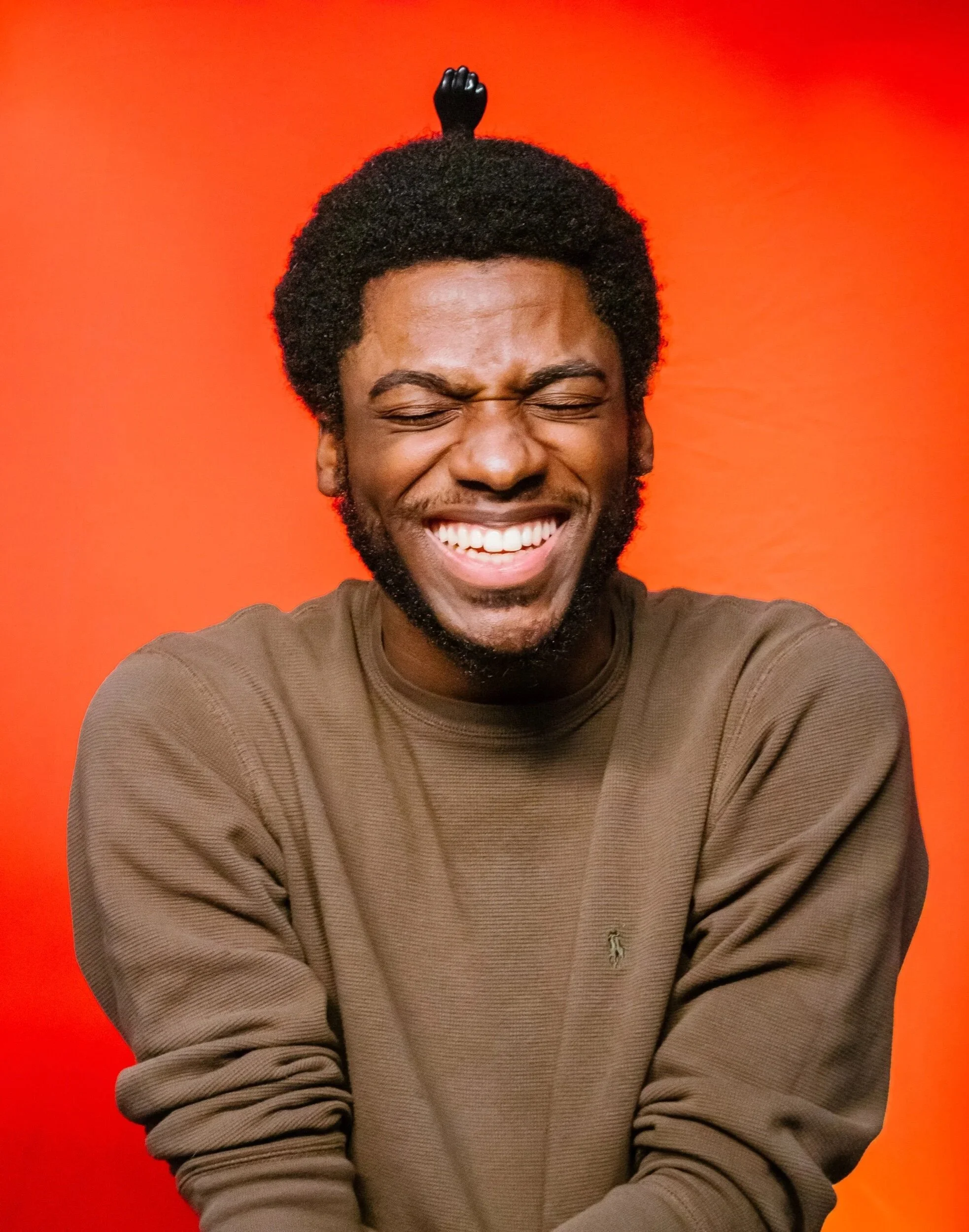 A man with curly black hair and a beard, laughing with his eyes closed, wearing a brown sweatshirt, against an orange background, with a small black figurine on top of his head.