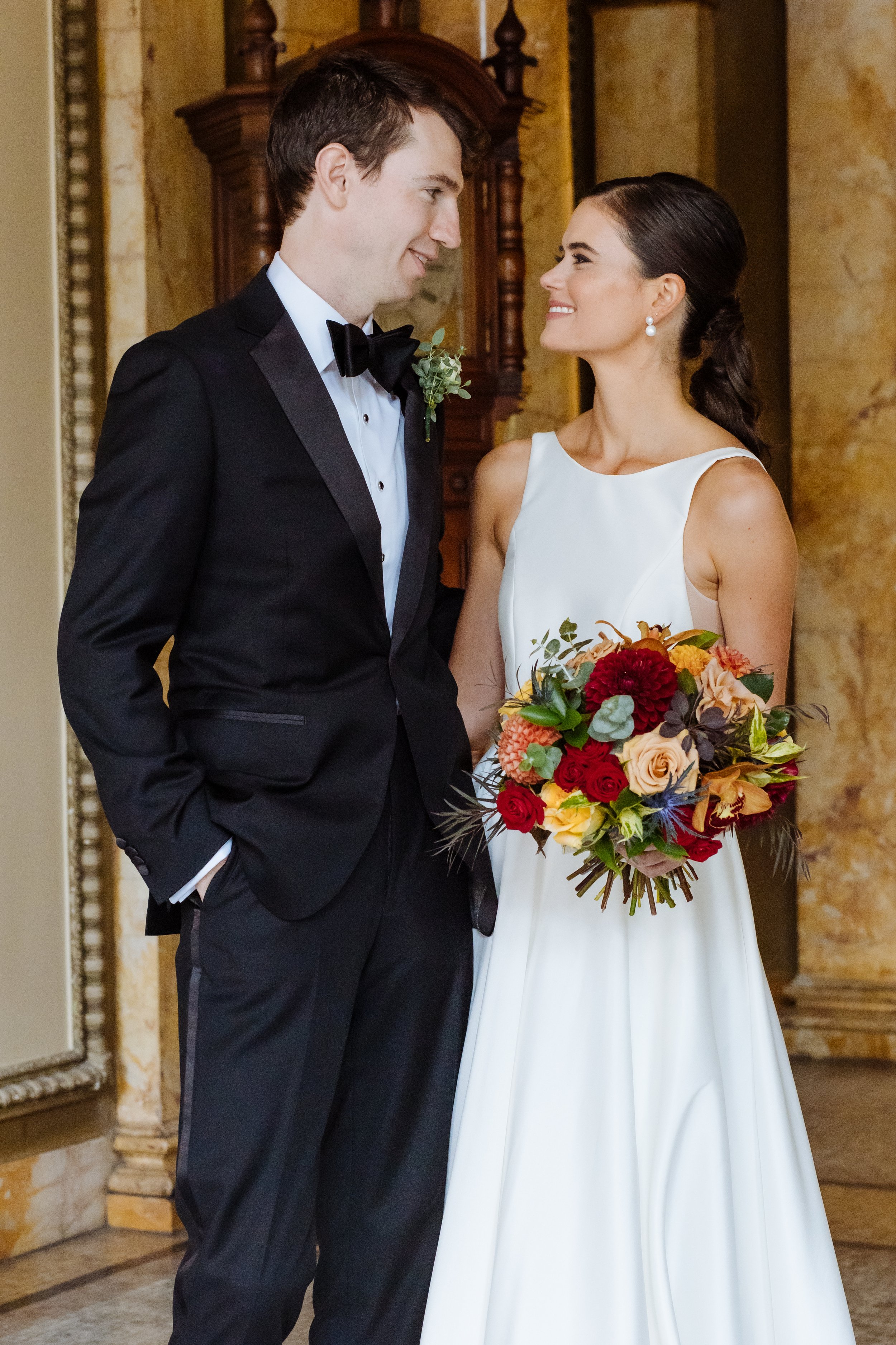 A bride and groom standing together in wedding attire, smiling at each other, indoors with a vintage style background. The bride is holding a colorful bouquet of flowers.