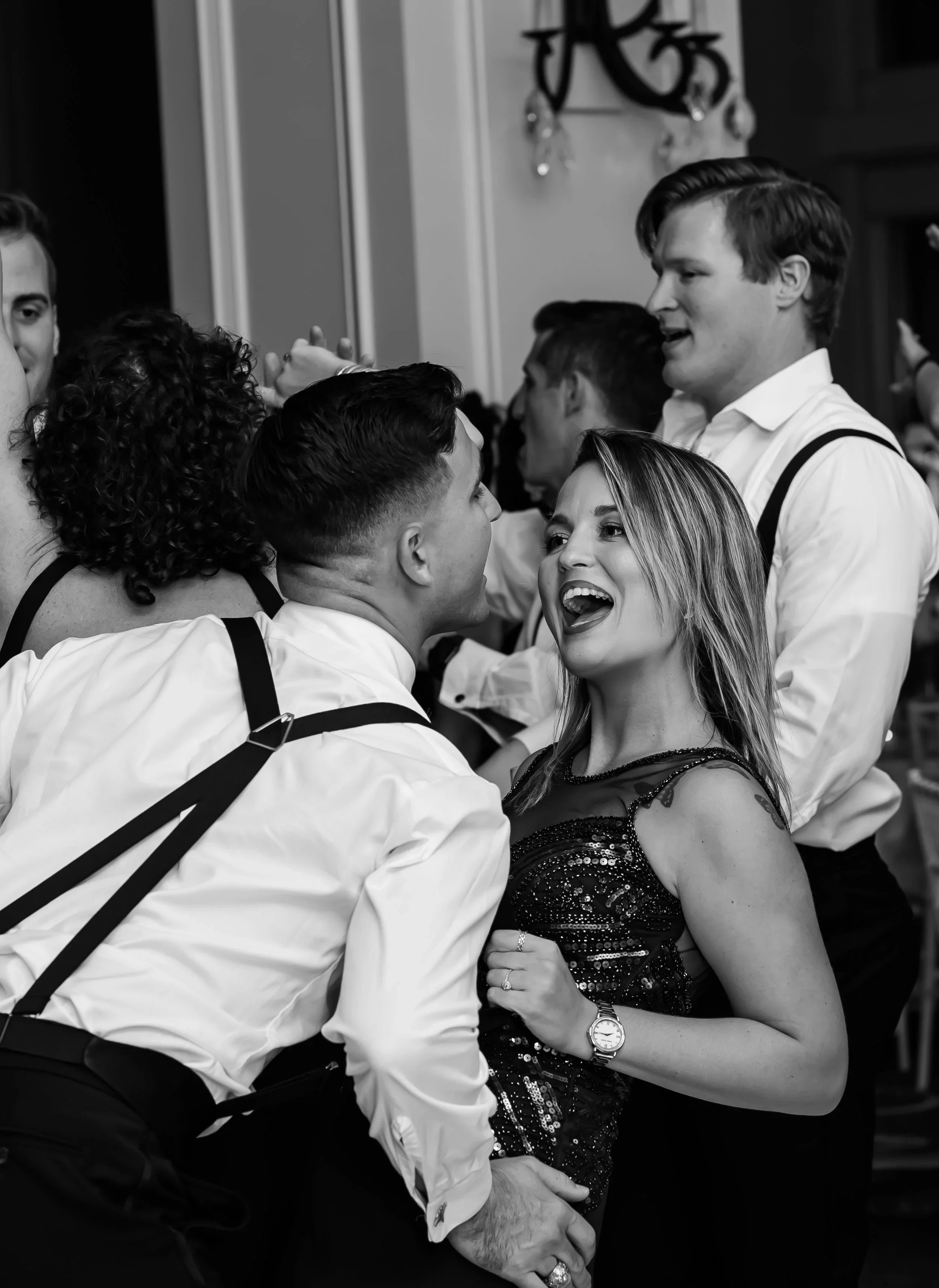 People dancing at a party, with a woman in a black sequined dress joyfully interacting with a man in a white shirt, surrounded by other dancers in formal attire.