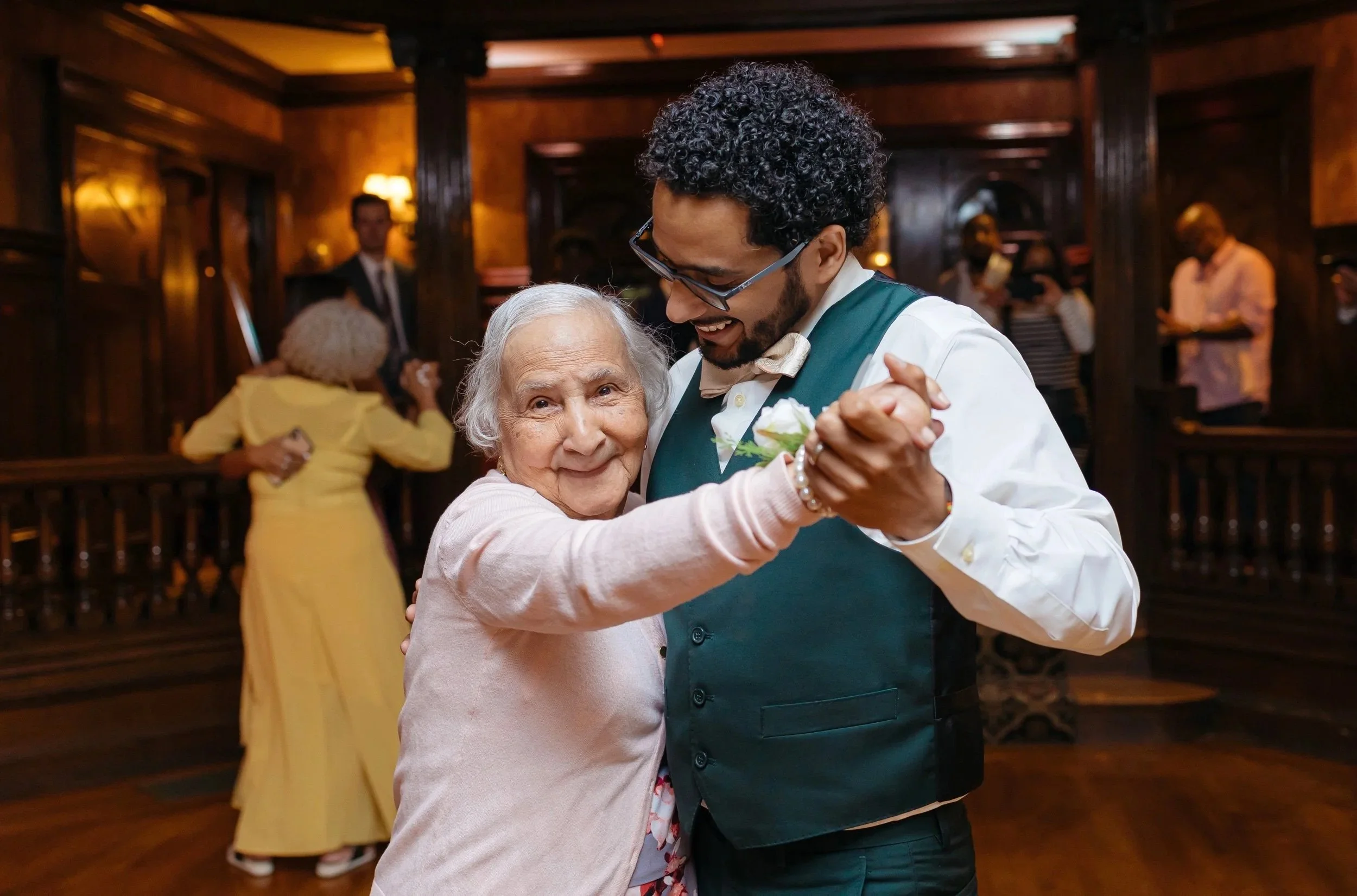 An elderly woman and a young man are dancing together at a social event in a warmly lit, wood-paneled room. The woman has gray hair and is wearing a light pink cardigan, while the man has curly black hair, glasses, and is dressed in a white shirt wit