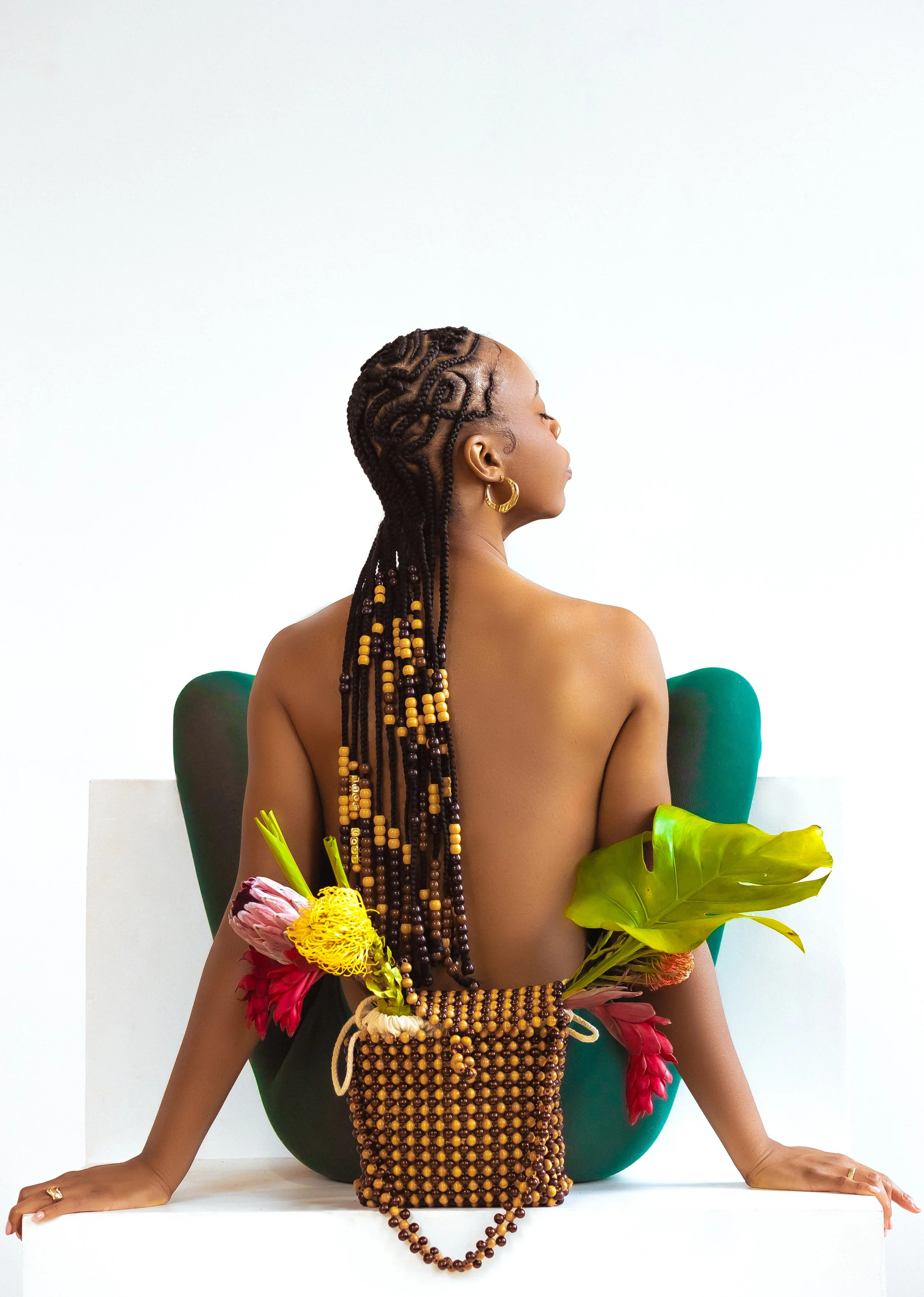 A woman with braided hair and gold hoop earrings is sitting with her back to the camera. She is surrounded by plants and flowers, with a beaded bag and necklace, and is seated on a white platform with a white background.