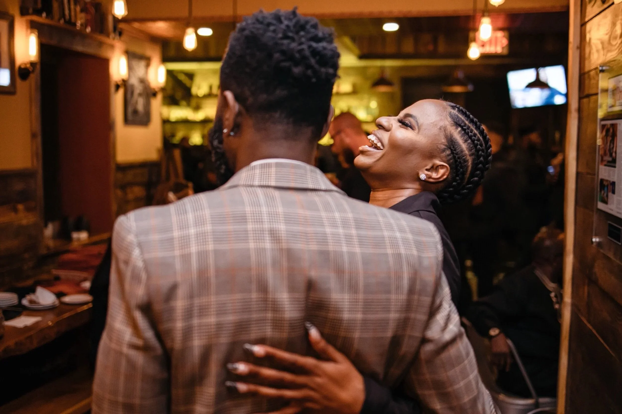 A woman with braided hair laughing and embracing a man with short curly hair in a restaurant or bar with warm lighting and wooden decor.