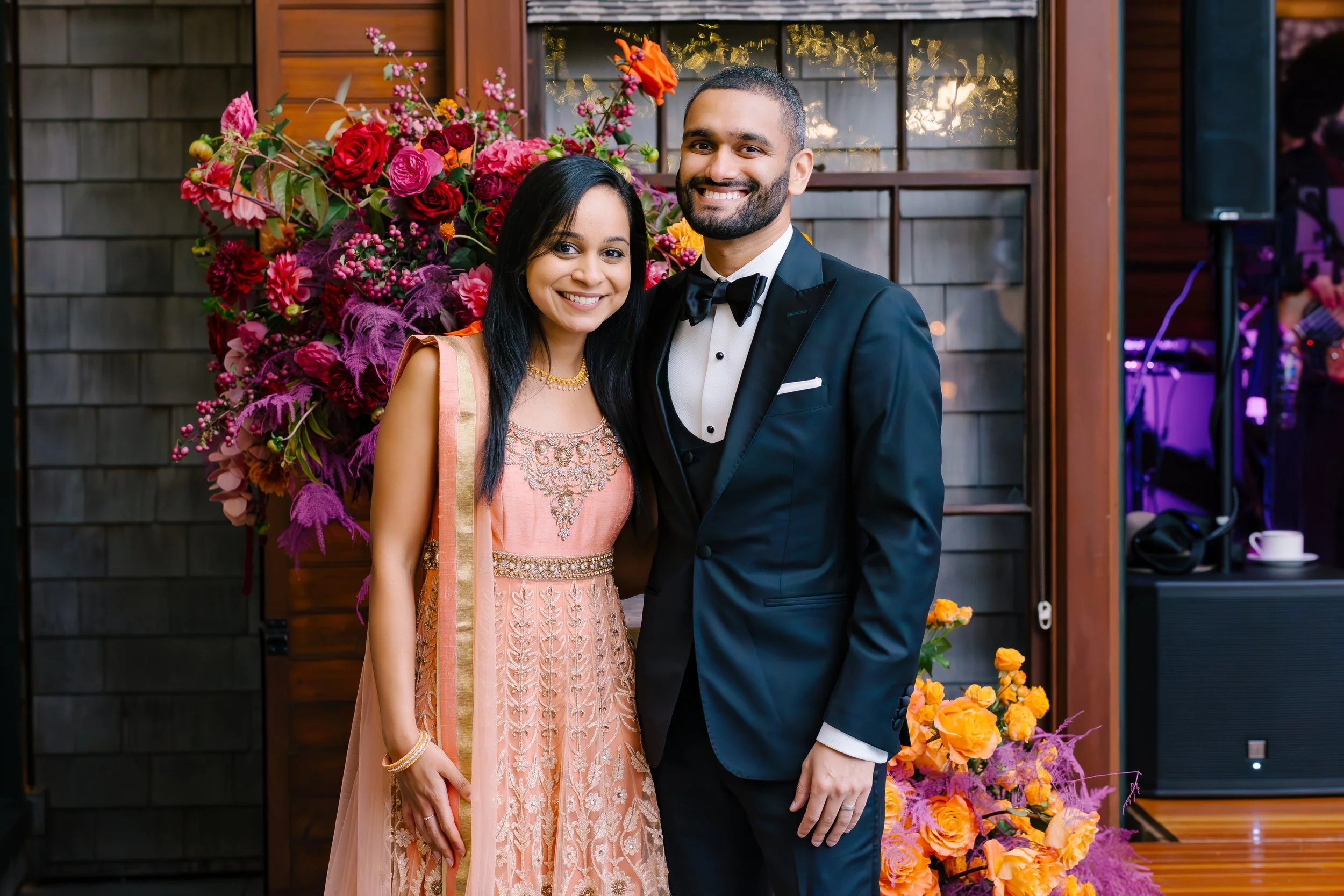 A couple dressed in formal attire, standing in front of a colorful floral arrangement at a celebration.