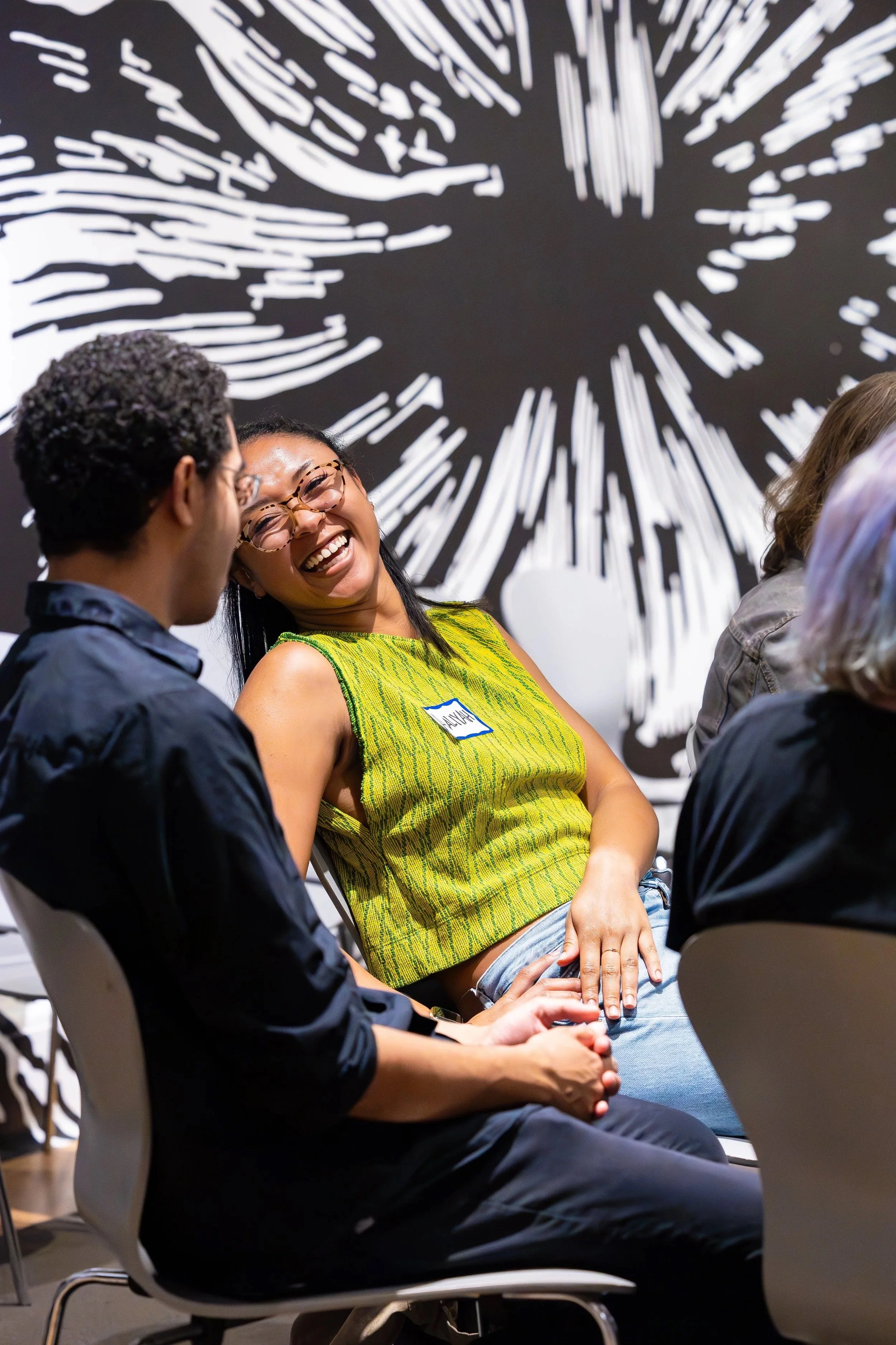 Two people sitting and talking, laughing, and smiling, with a black and white abstract wall as background.