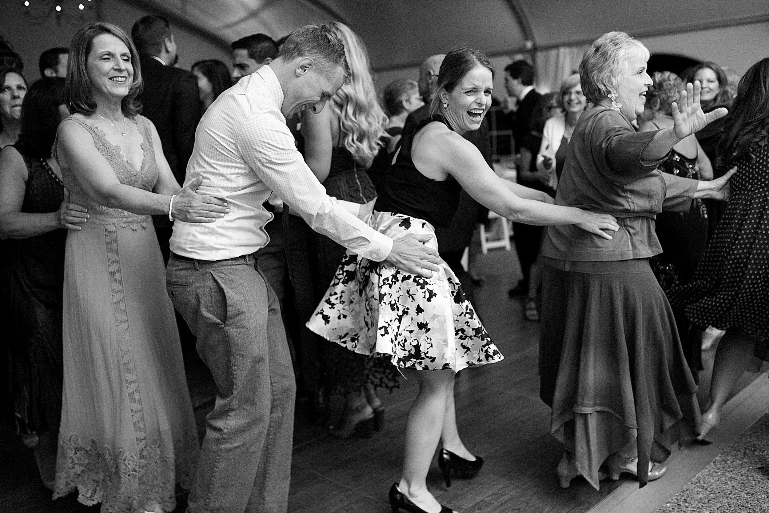 Group of people dancing at a party, holding onto each other's waists in a conga line, smiling and enjoying themselves.