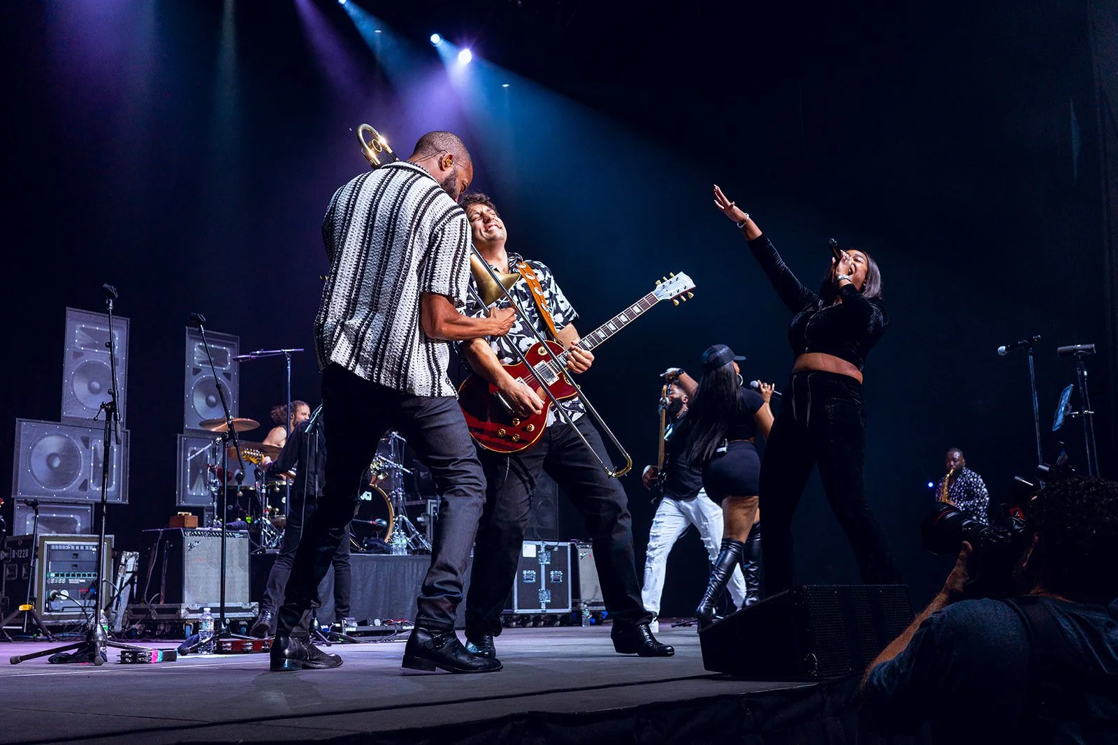 A live musical performance with Trombone Shorty and his band on stage, with purple and blue stage lighting.