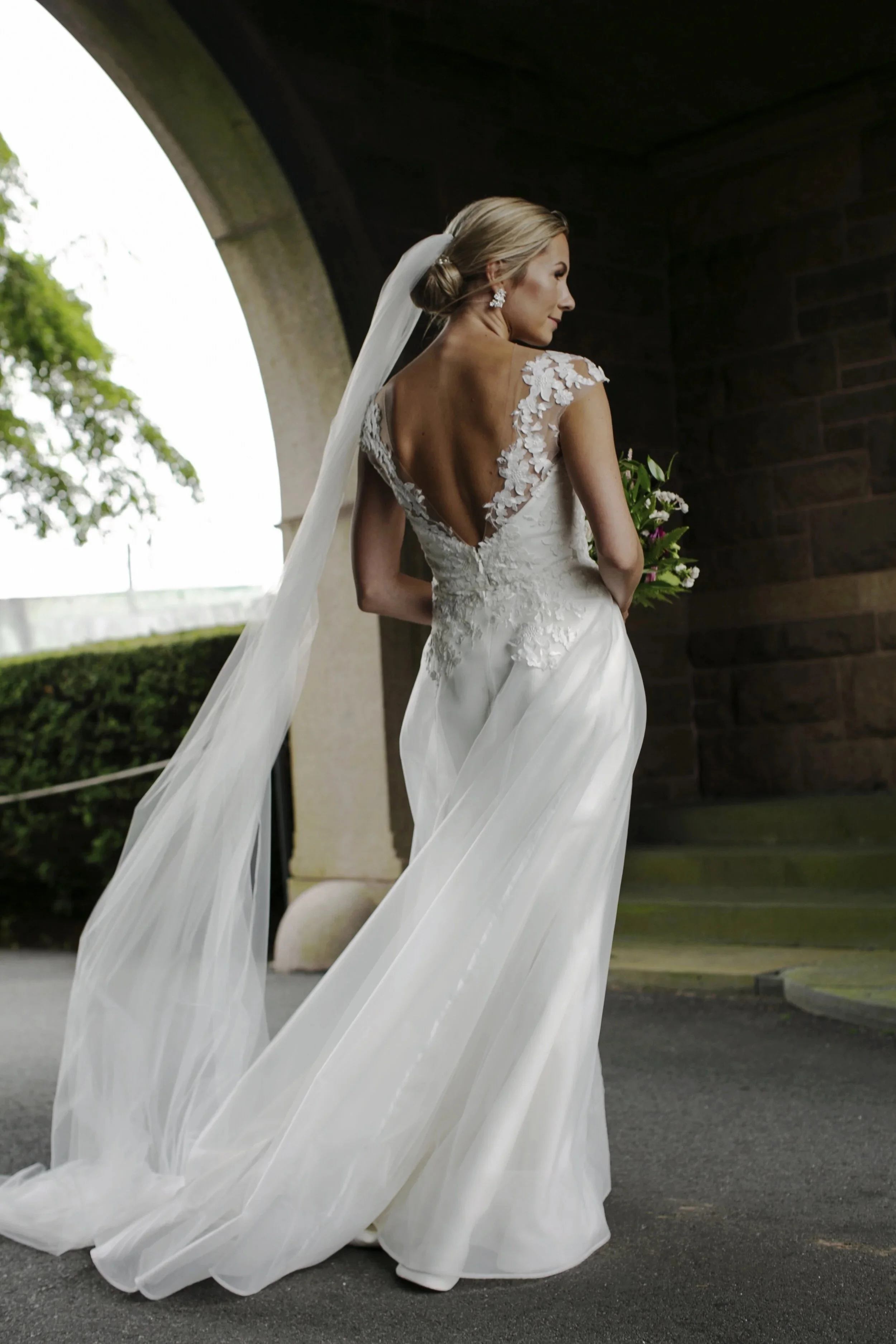 Bride in a white wedding gown with lace details, holding a bouquet, standing under an arched bridge, turned to the side.