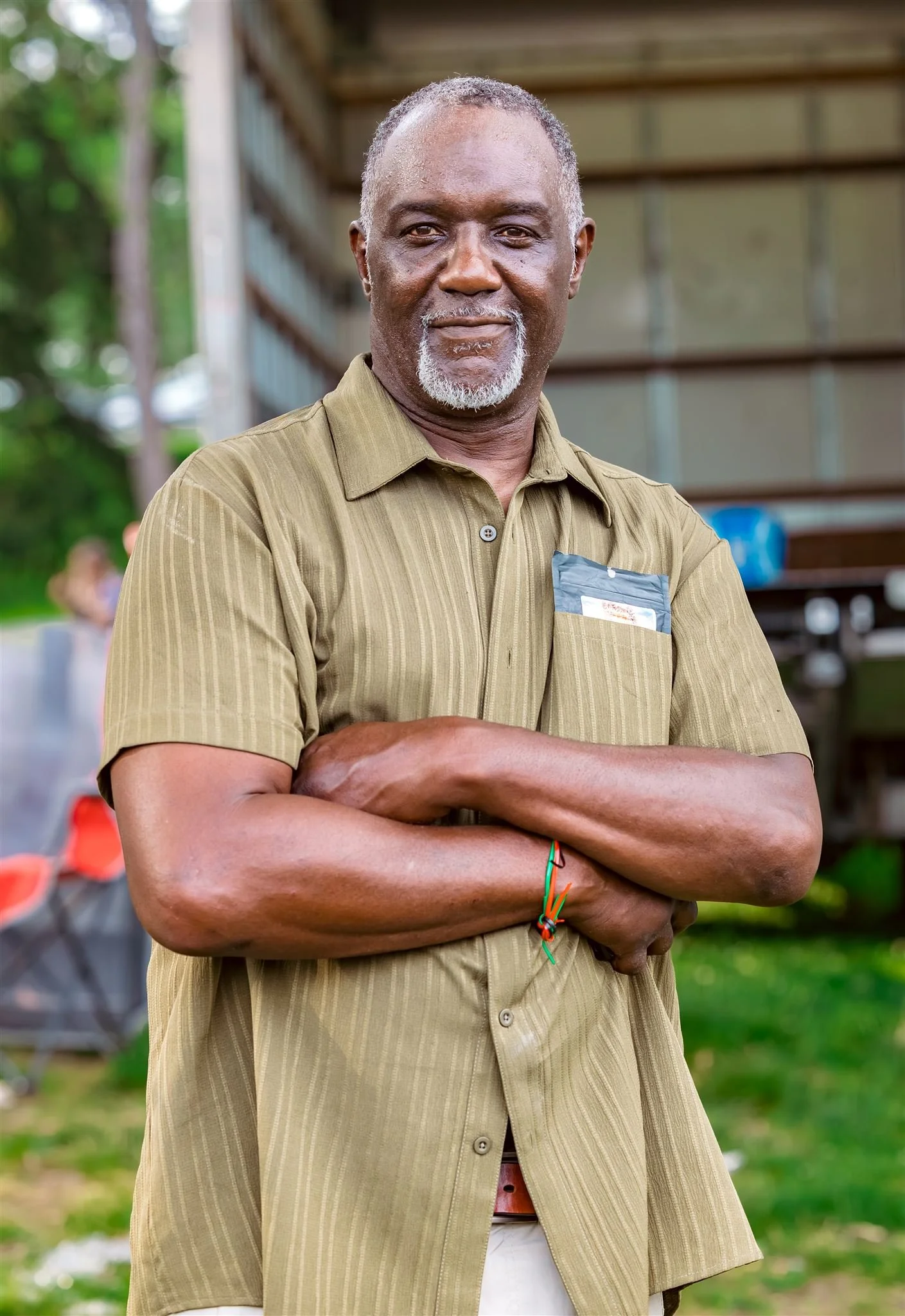 A smiling older man with gray hair and beard, wearing a light brown striped shirt with his arms crossed, standing outdoors in a garden or park area.