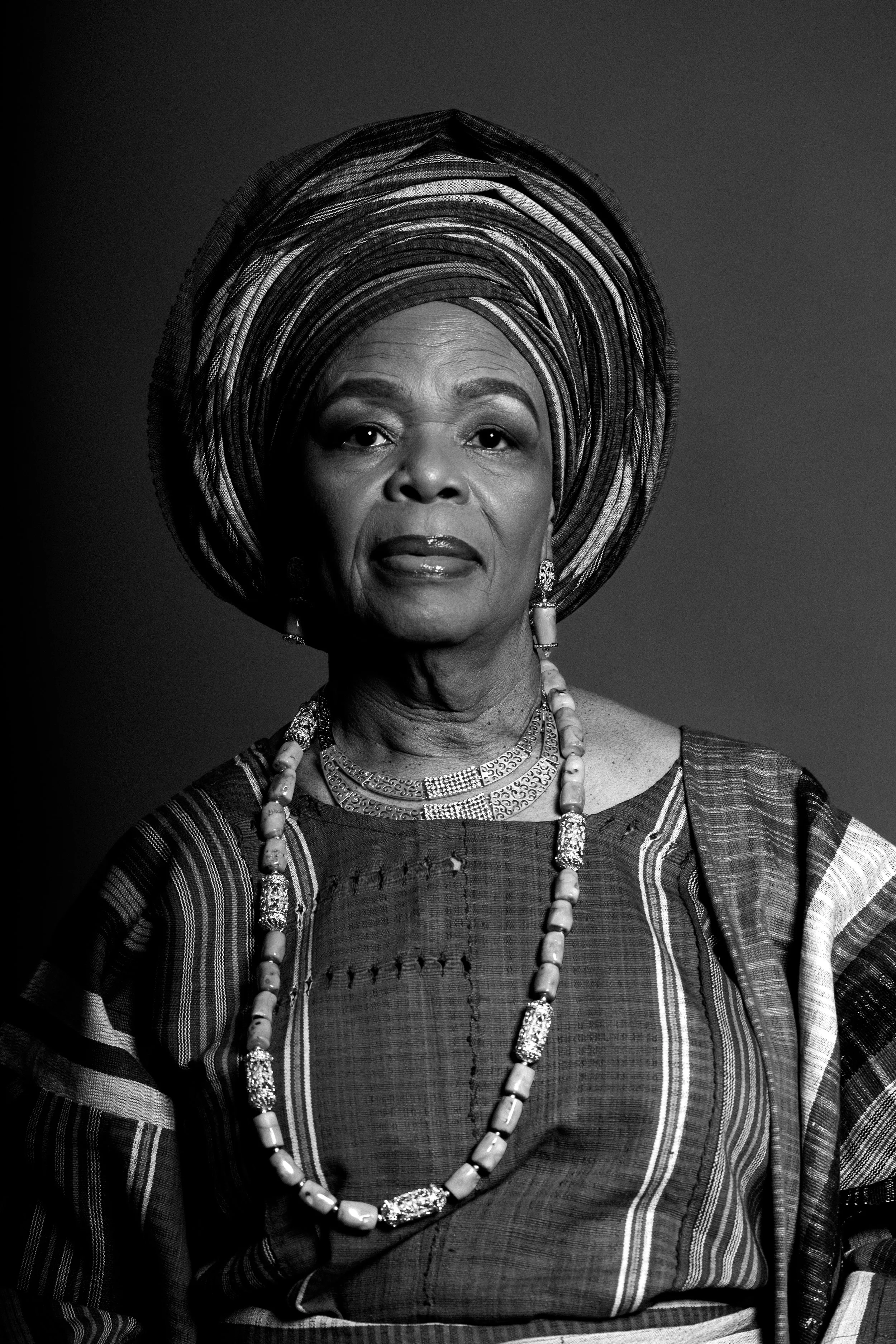 A woman wearing traditional African clothing and headwrap, with jewelry, in a black-and-white portrait.