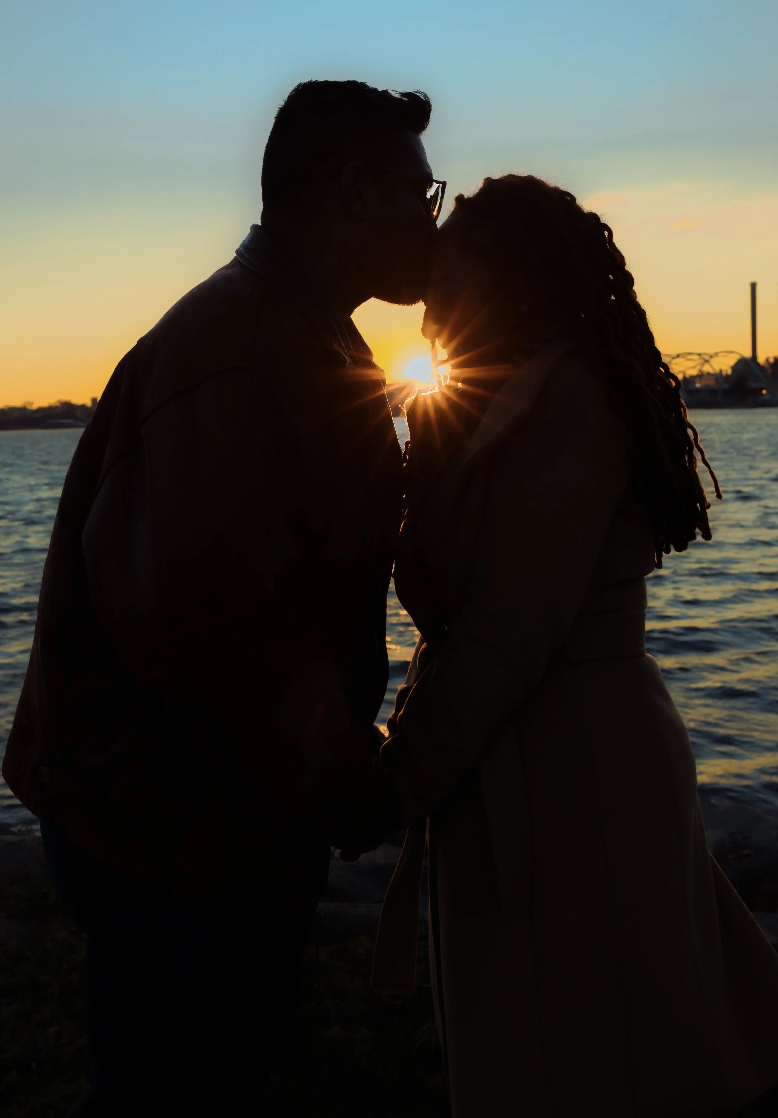 A couple standing by the water at sunset, leaning in to kiss, with the sun setting behind them.