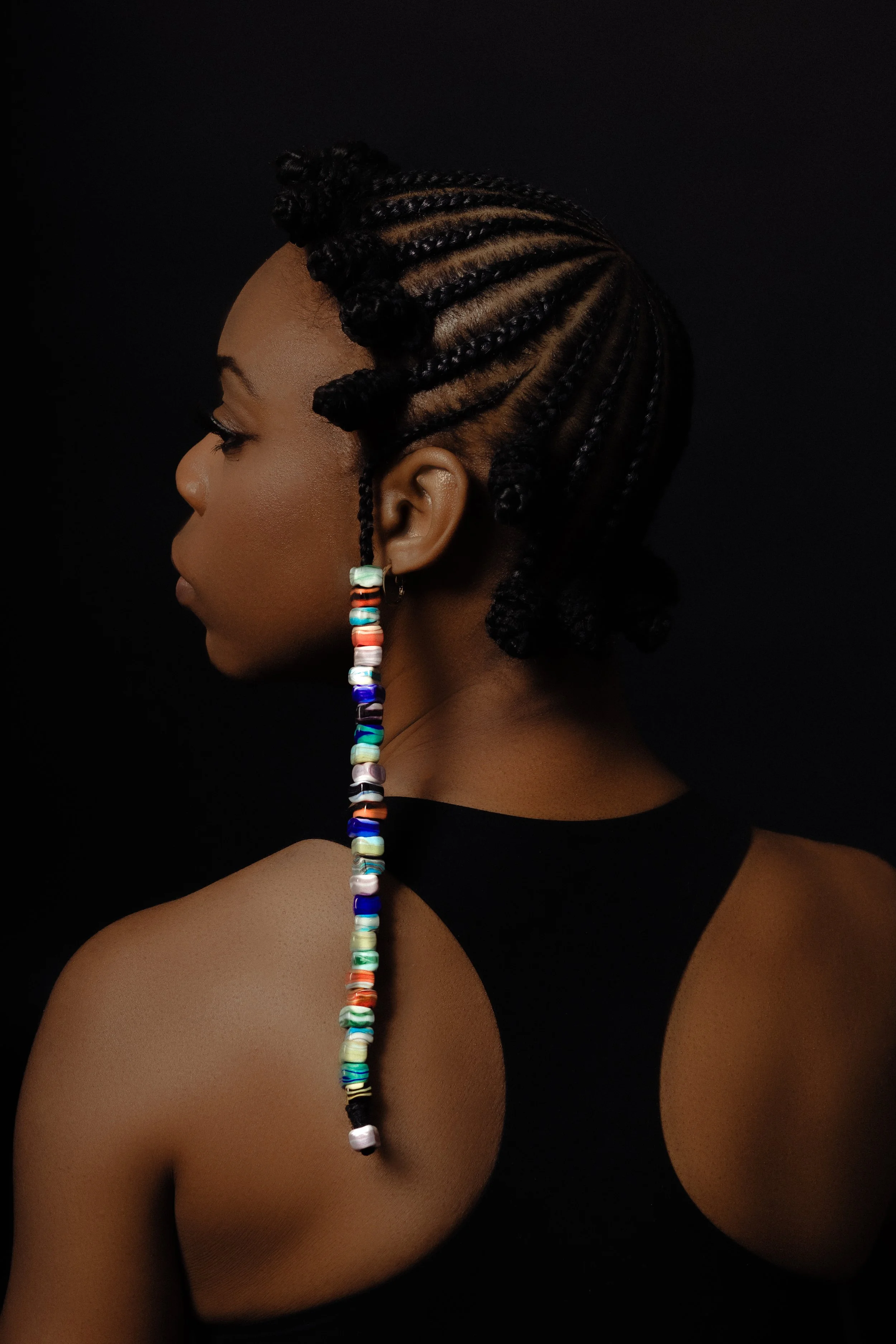 A woman with styled cornrow hair and a colorful beaded earring, wearing a black sleeveless top, against a dark background.