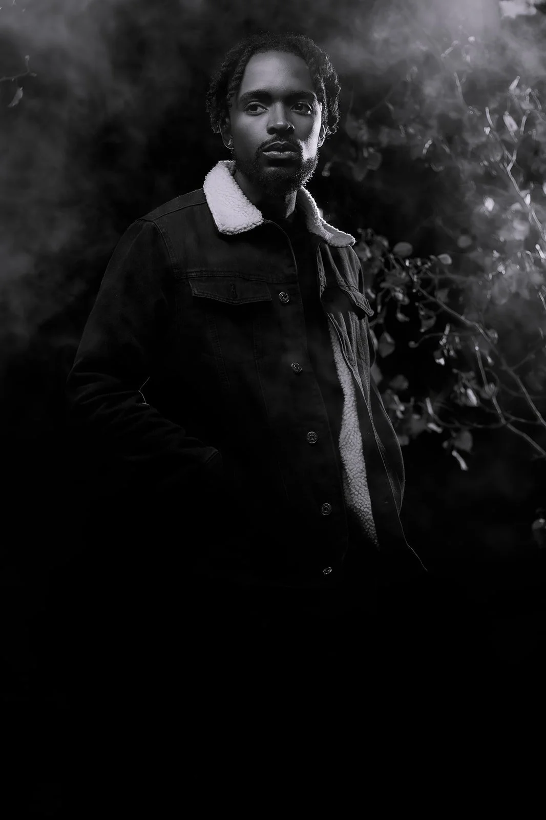 A black and white portrait of a man with dreadlocks, wearing a jacket with a shearling collar, standing outdoors near some foliage.