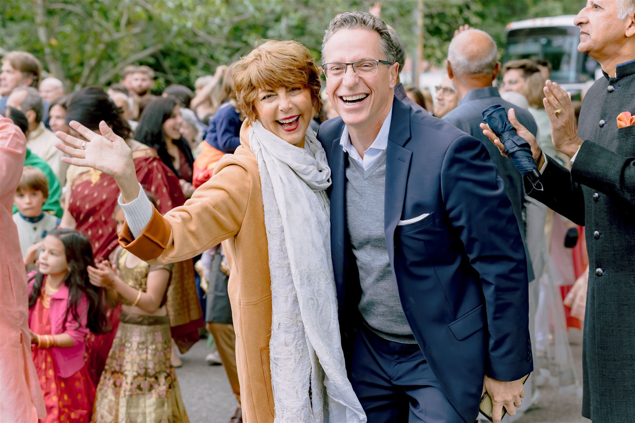 A joyful gathering at an outdoor event with a woman in a tan coat and white scarf, and a man in a navy blazer, smiling and celebrating among a crowd of diverse people, including children and seniors.