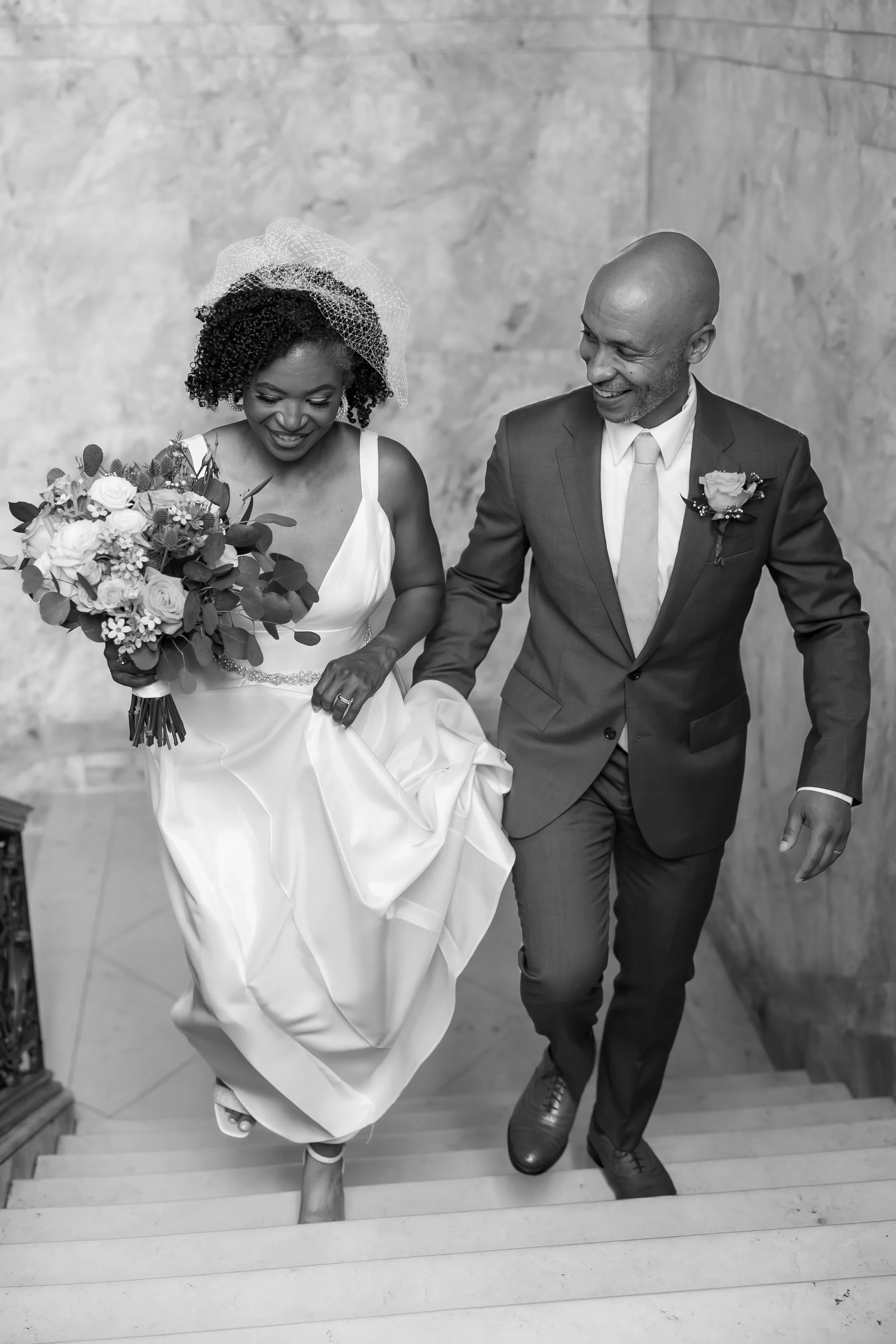 A bride and groom walking up stairs, smiling, with the bride holding a bouquet of flowers, during a wedding ceremony.