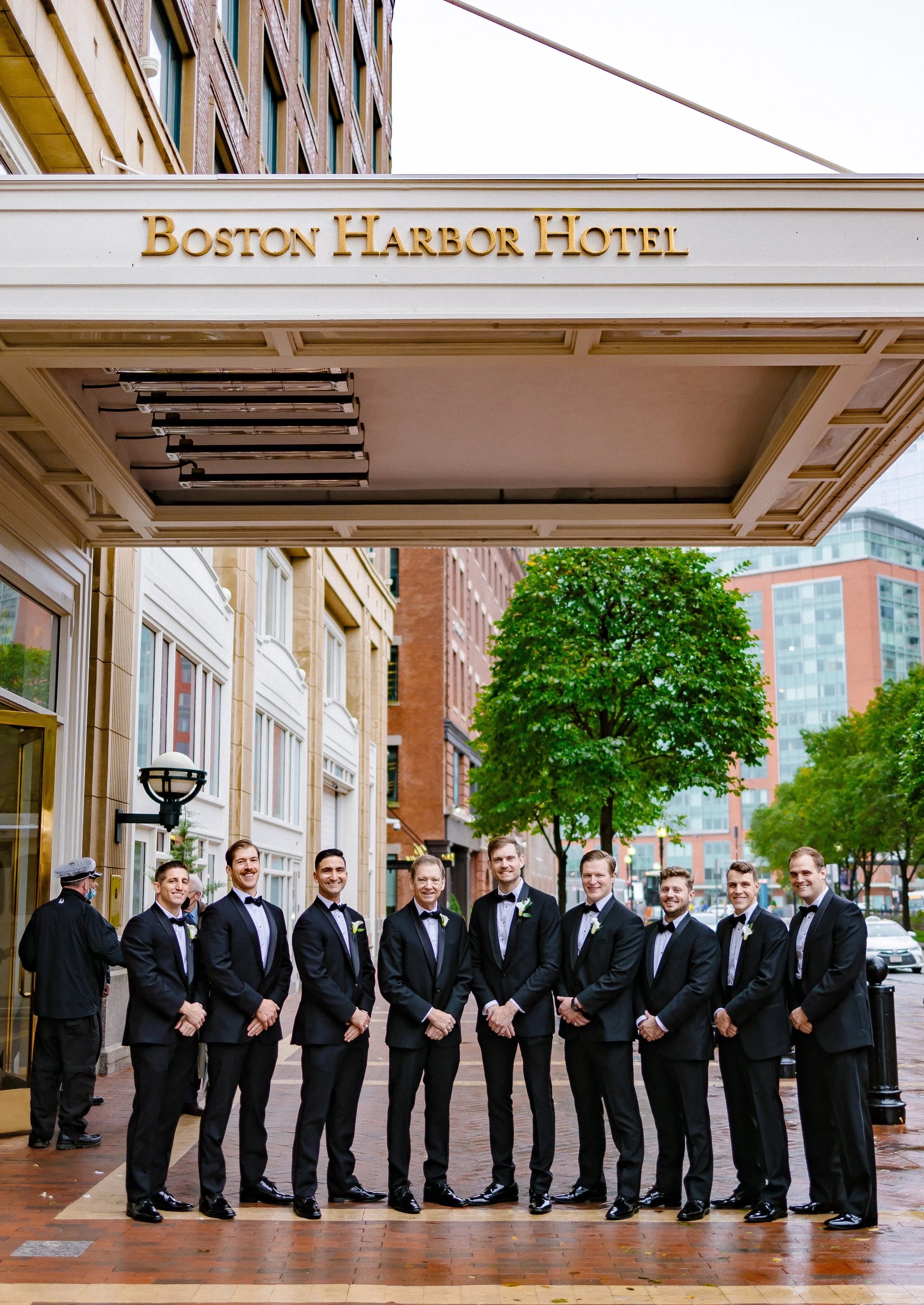 A group of eight men dressed in black tuxedos with bow ties standing outside the Boston Harbor Hotel. Behind them are city buildings and green trees, and the hotel entrance is visible above.