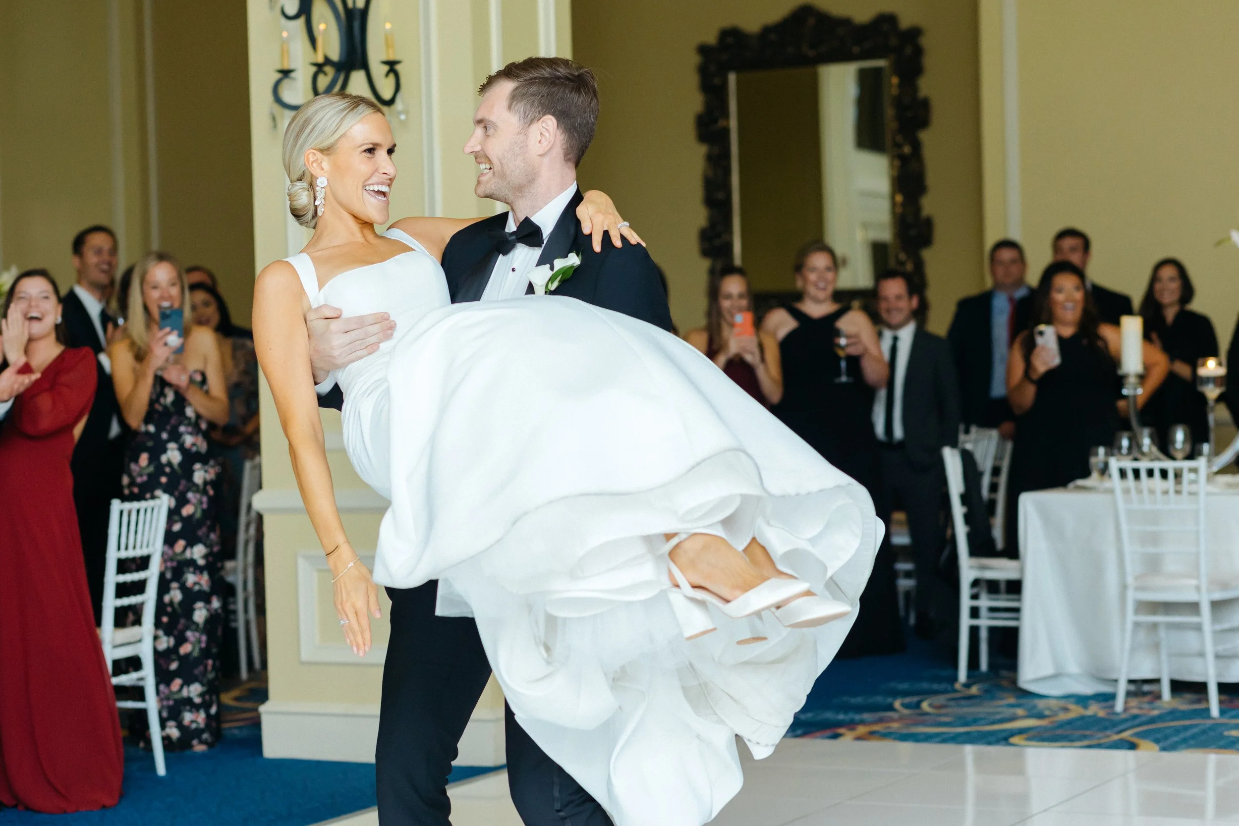 A groom in a black tuxedo carries a smiling bride in a white wedding dress during their wedding reception. Guests in formal attire watch and smile, some taking photos, in a decorated banquet hall with lights, candles, and a large mirror in the backgr