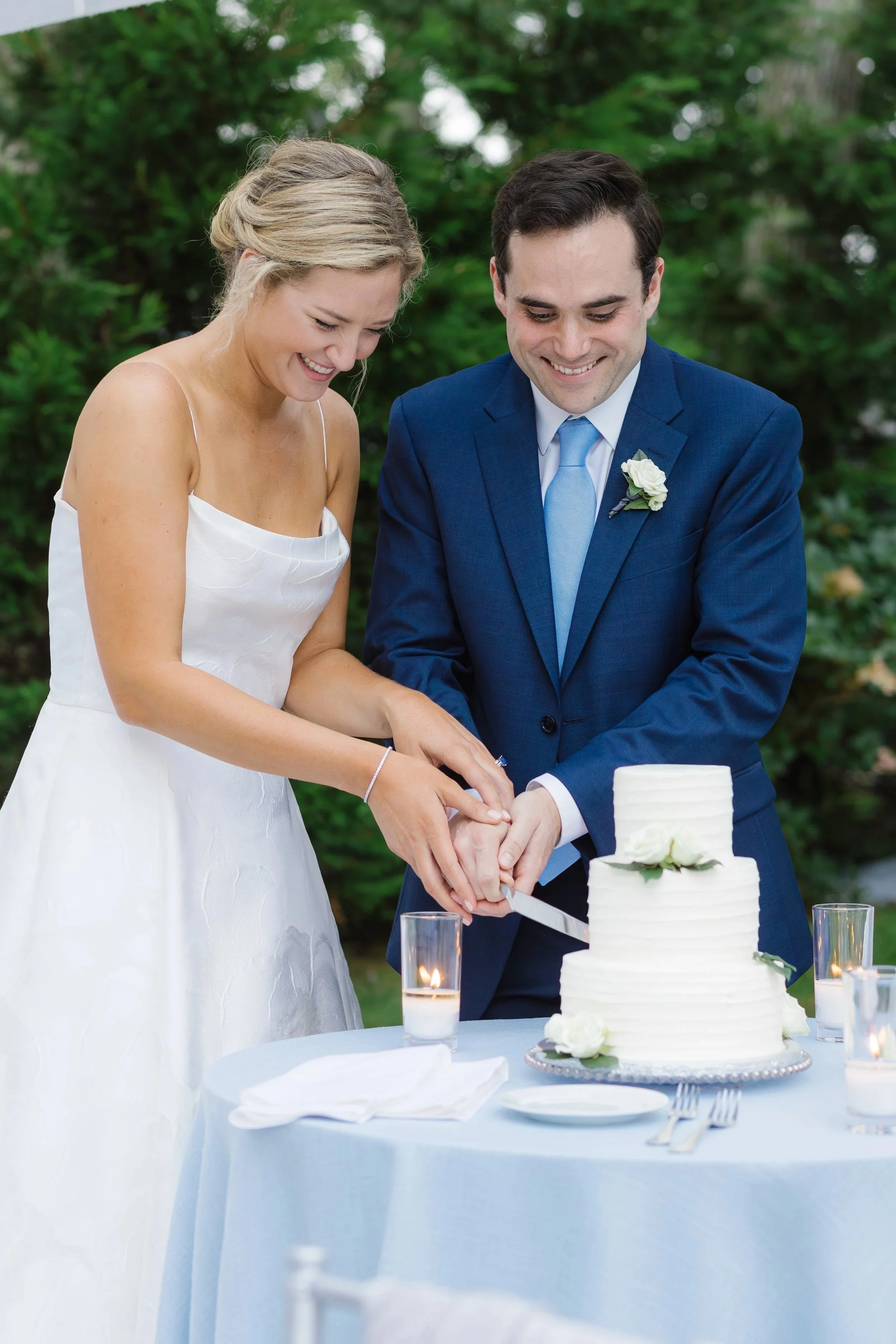 A newlywed couple in wedding attire cuts a wedding cake at an outdoor reception, with greenery in the background and lit candles on the table.