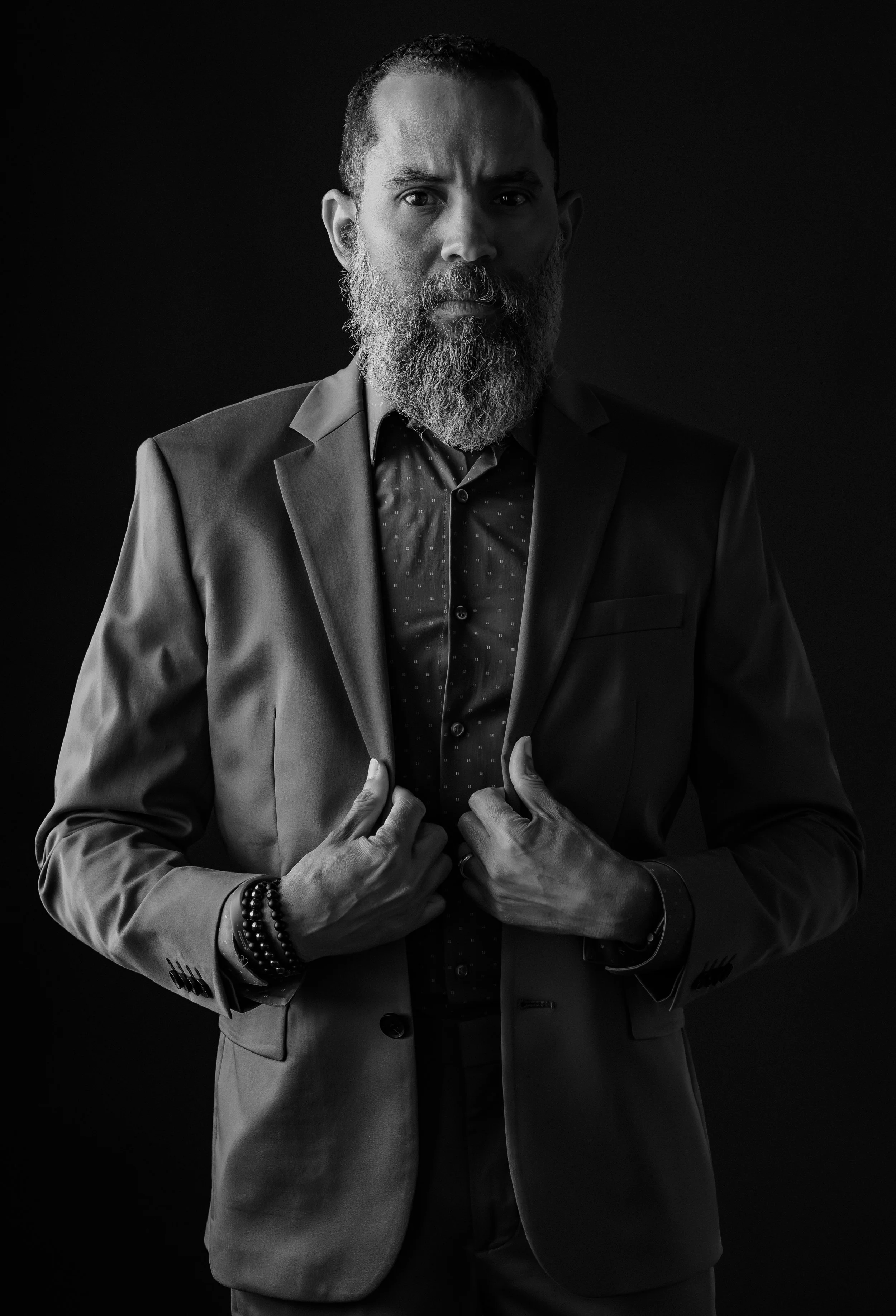 Black and white portrait of a man with a beard wearing a suit jacket and a patterned shirt, adjusting the jacket with both hands, against a dark background.