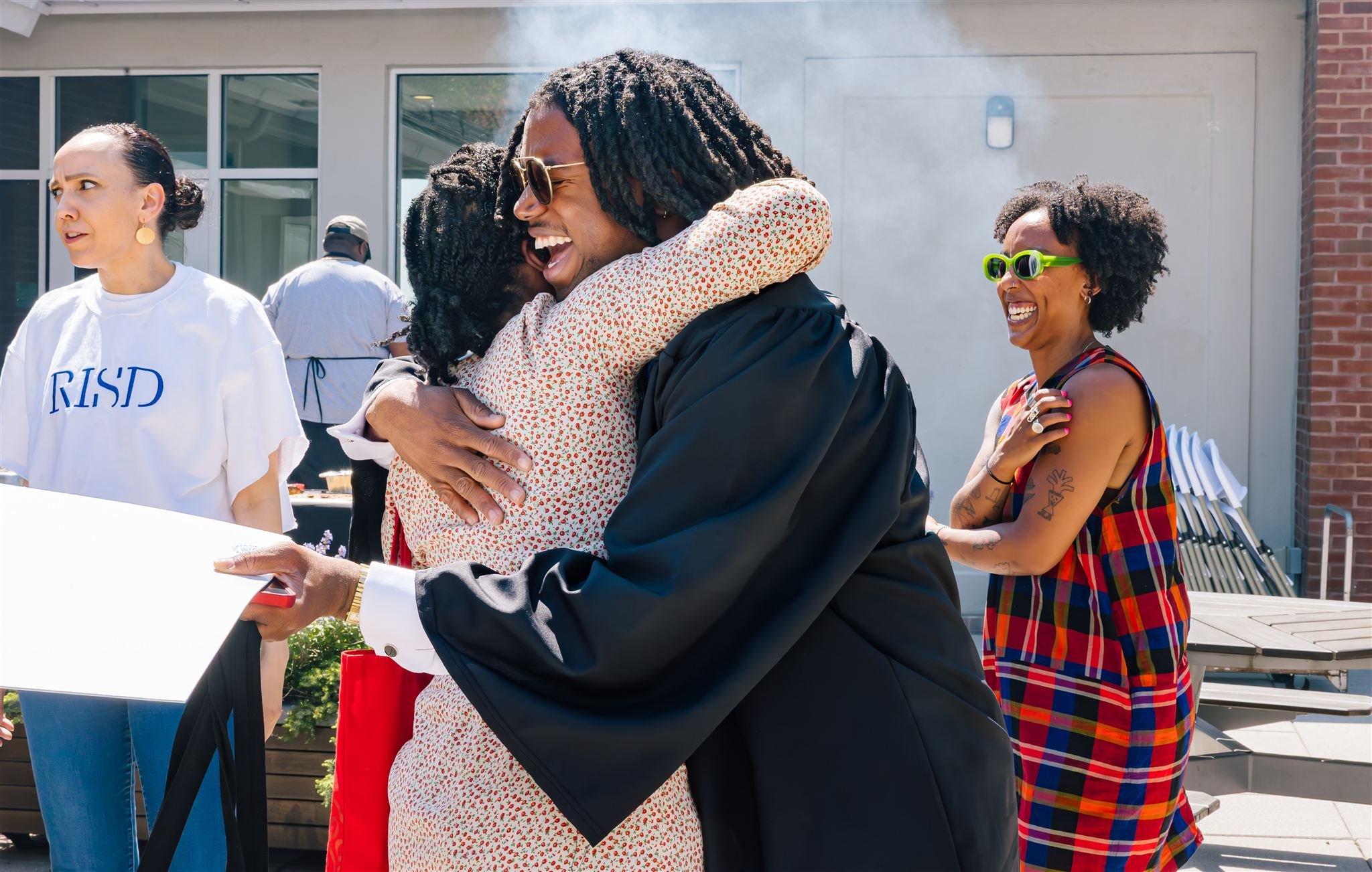 Group of people celebrating outdoors, hugging and smiling, with one woman in sunglasses and a plaid dress, and others in casual clothing.