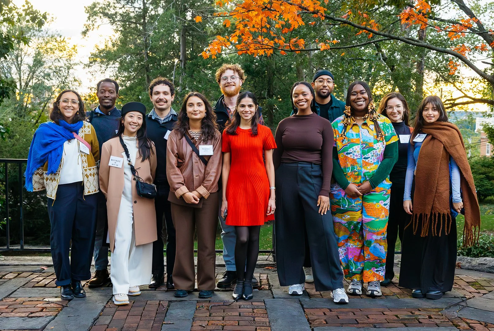 A diverse group of fifteen people standing outdoors on a brick path with trees in the background, smiling at the camera.