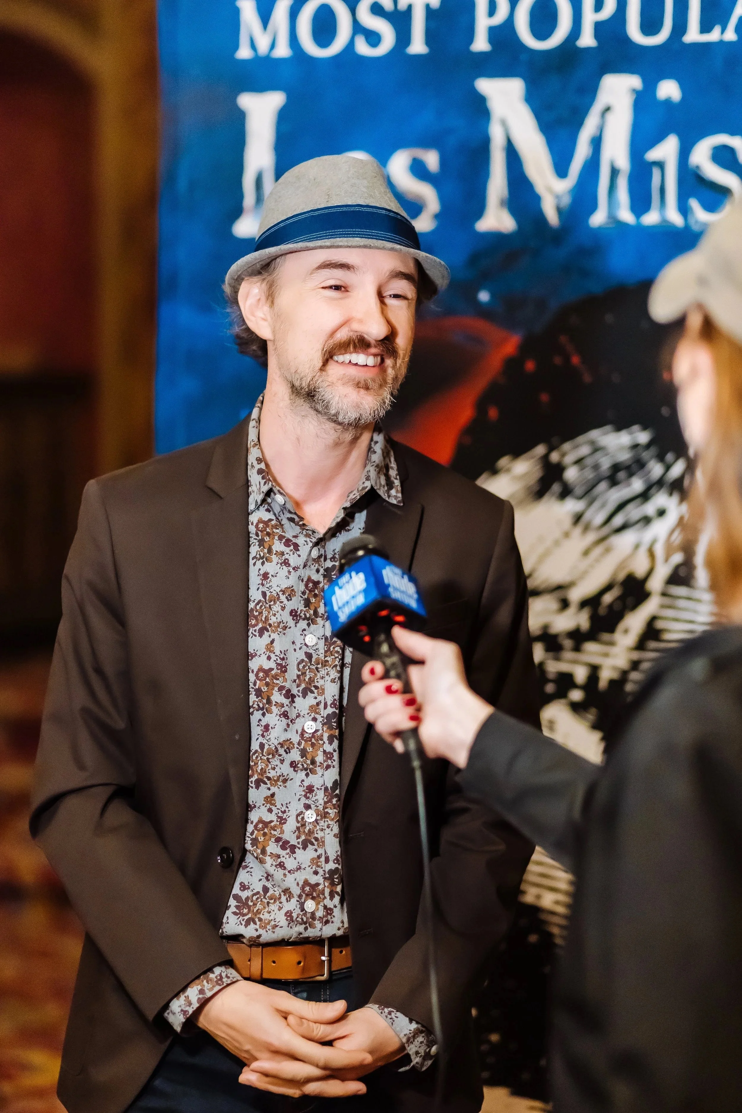 A man with a grey hat, patterned shirt, and dark blazer being interviewed at an event with a blue banner that has white text and an image of a guitar in the background.