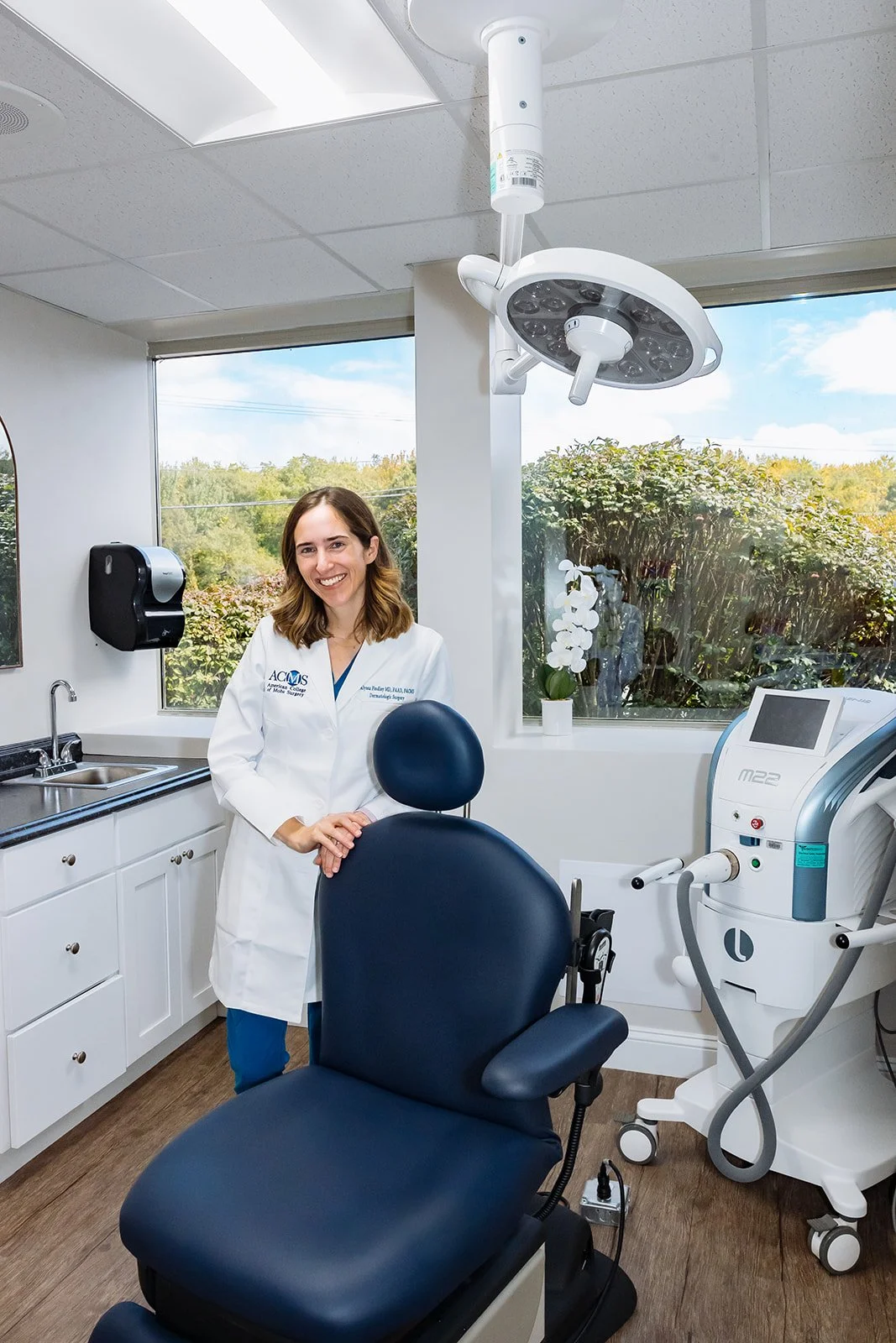 A smiling female healthcare professional in a white lab coat standing beside a dental or medical chair in a clean, well-lit clinic room, with medical equipment and a large window showing trees outside.