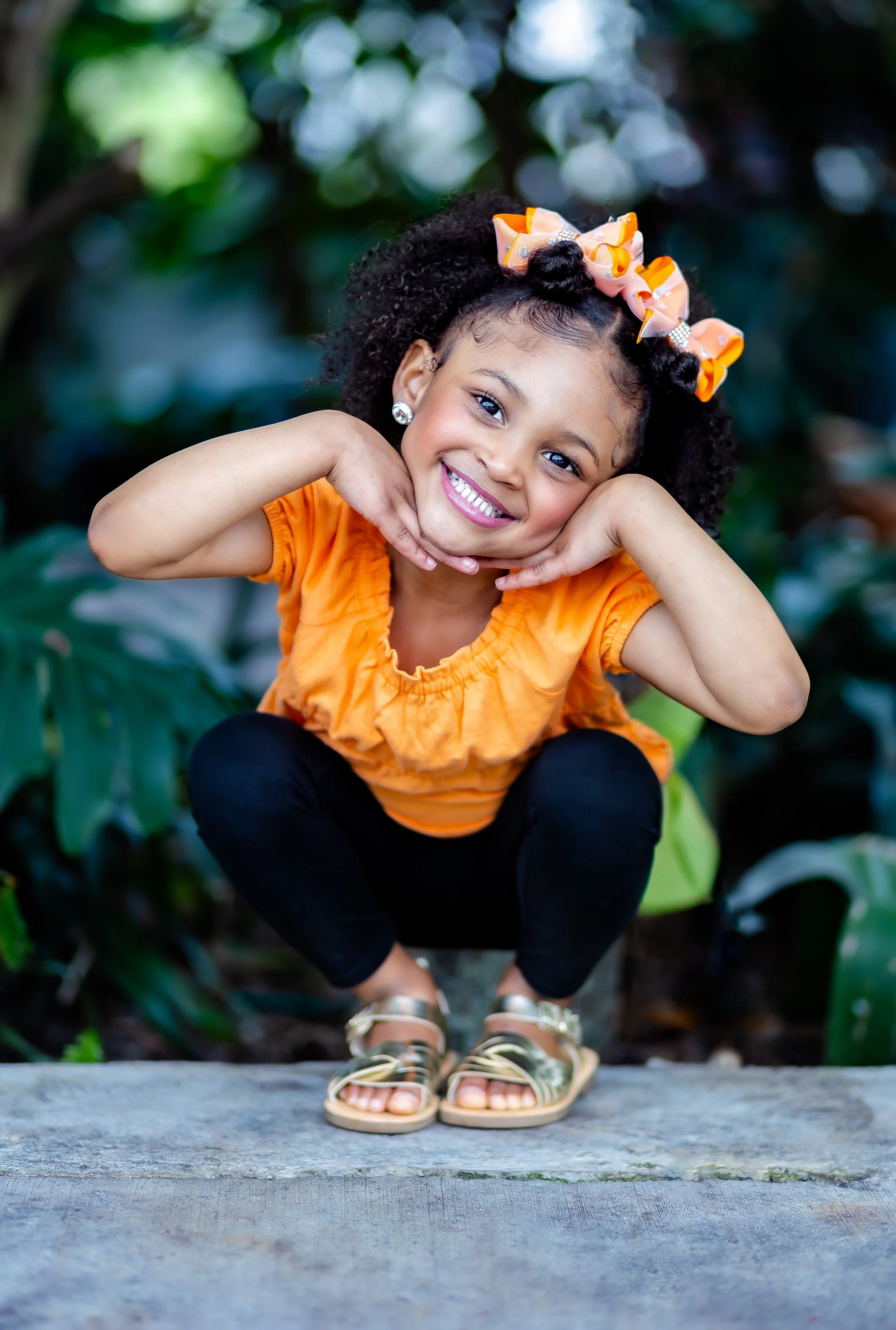 A young girl with curly hair, wearing an orange top, black pants, and gold sandals, crouches on a wooden surface and smiles with her hands under her chin, in a lush outdoor setting.